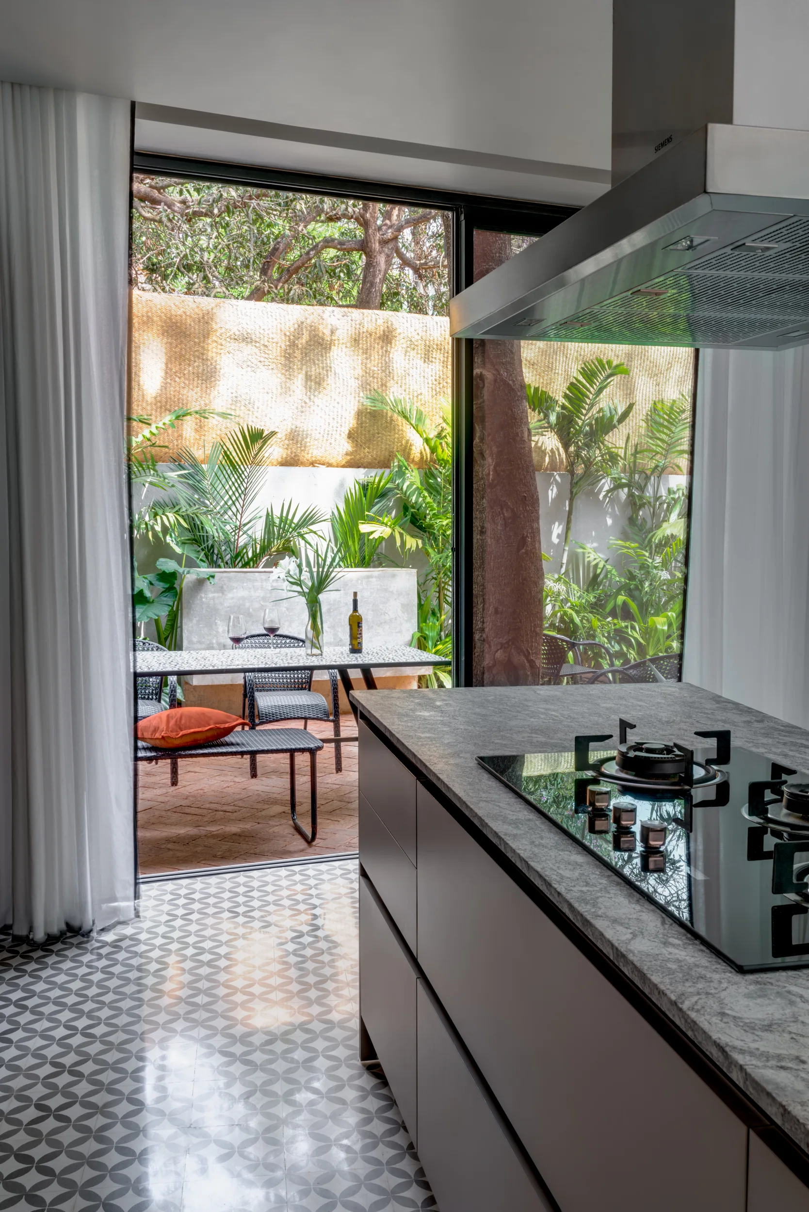 A modern kitchen island with a dark cooktop sits on patterned geometric floor tiles next to open sliding glass doors leading to a patio.