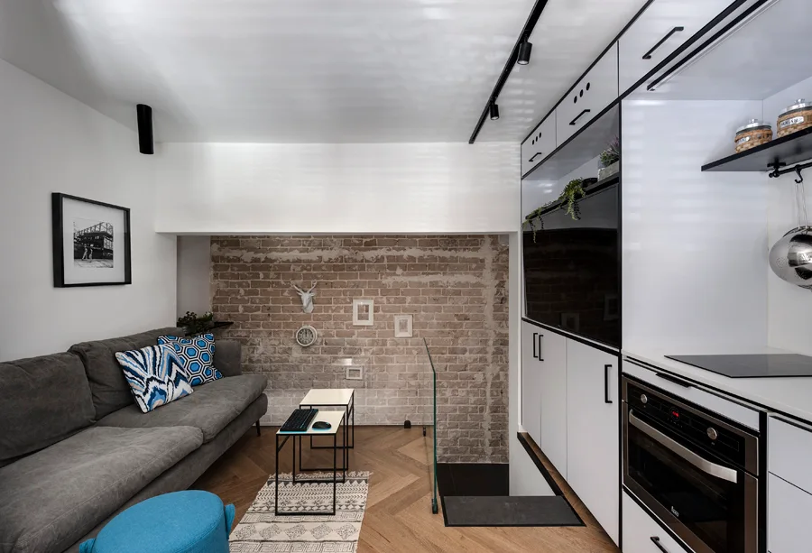 A compact living area features a grey sofa against an exposed brick wall, alongside sleek white kitchen cabinets and herringbone wood floors.