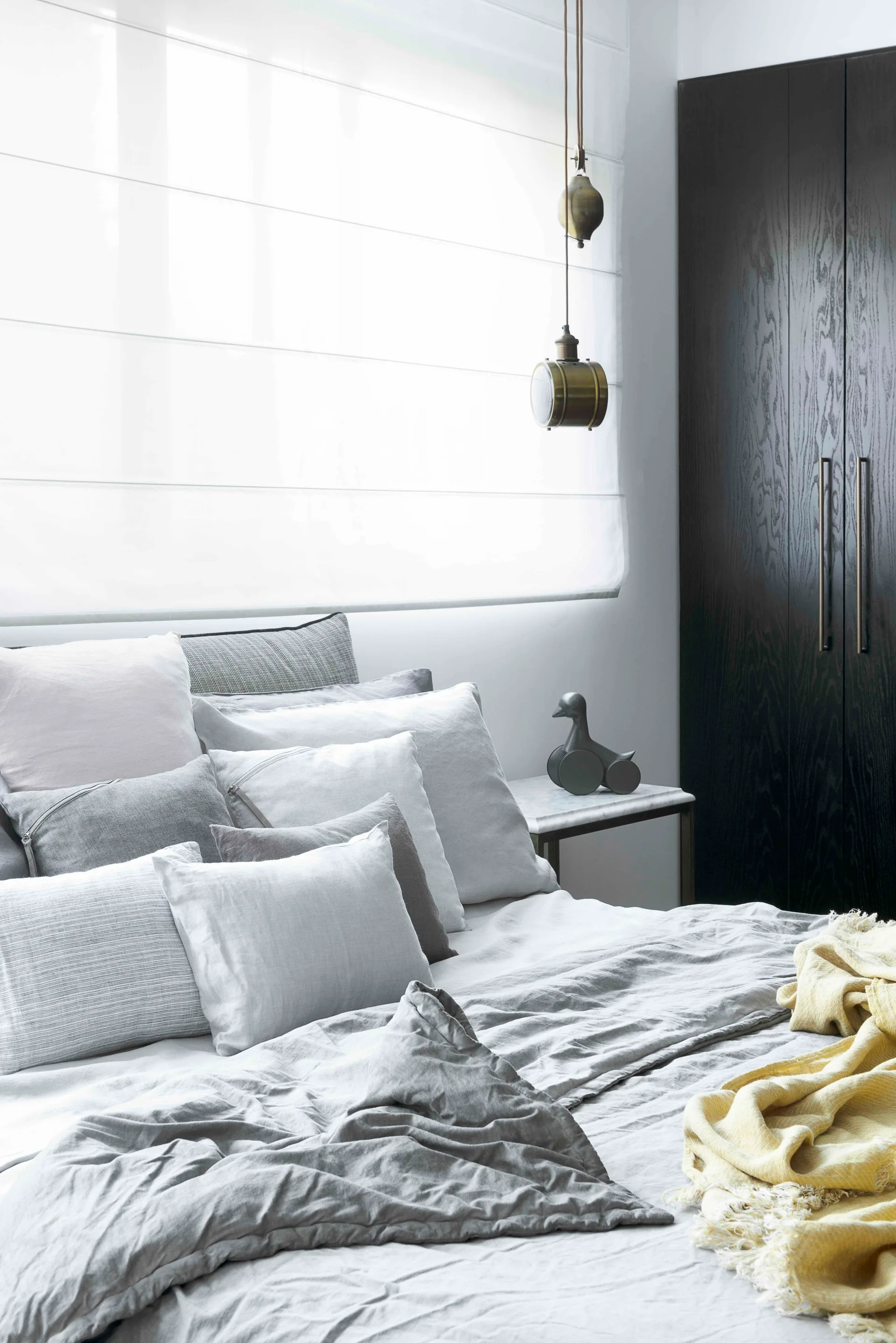 A modern bedroom features layered grey linen bedding, a white roman shade, a dark wood wardrobe, and a brass pendant light above a marble nightstand.