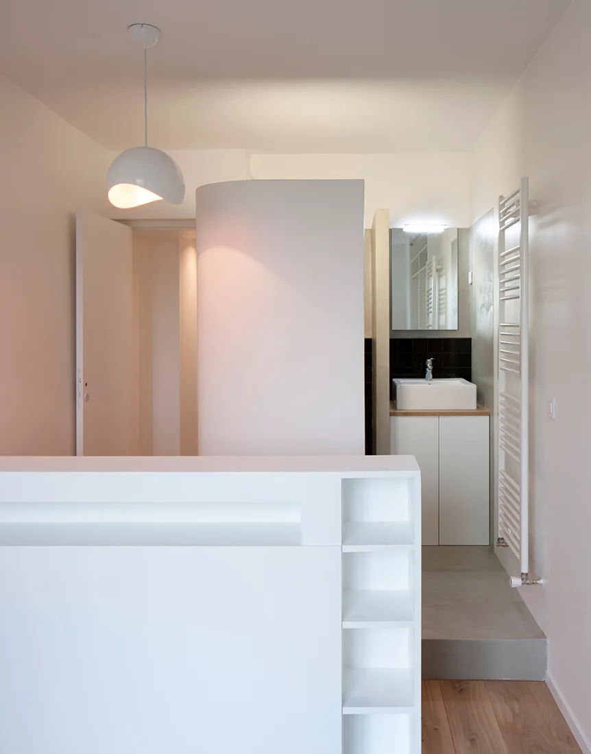 A minimalist white bathroom featuring a curved partition wall, integrated open shelving, a sink vanity on a concrete step, and a white towel warmer.