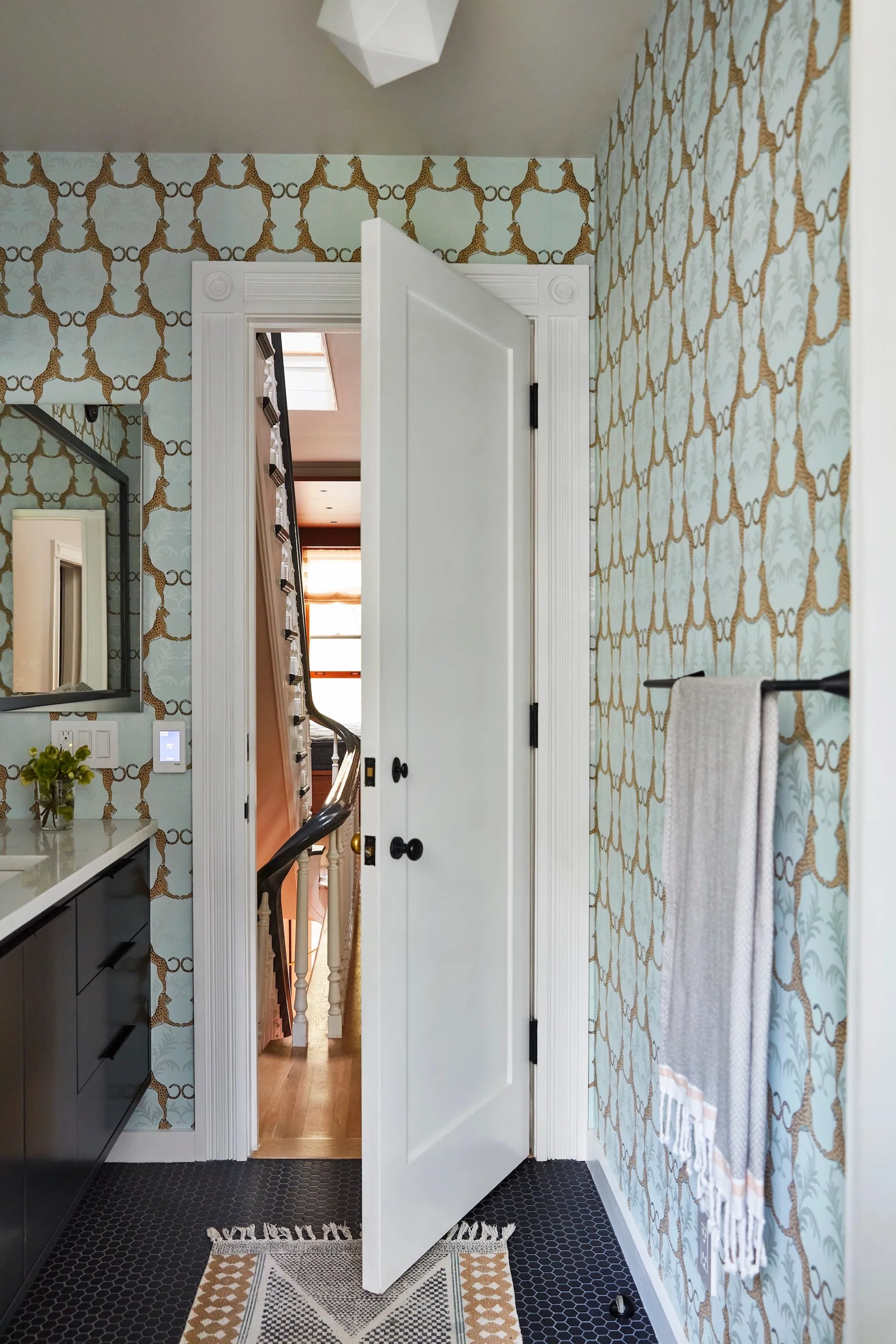 A powder room features light blue animal print wallpaper, a dark vanity, black hexagonal floor tiles, and an open white door.