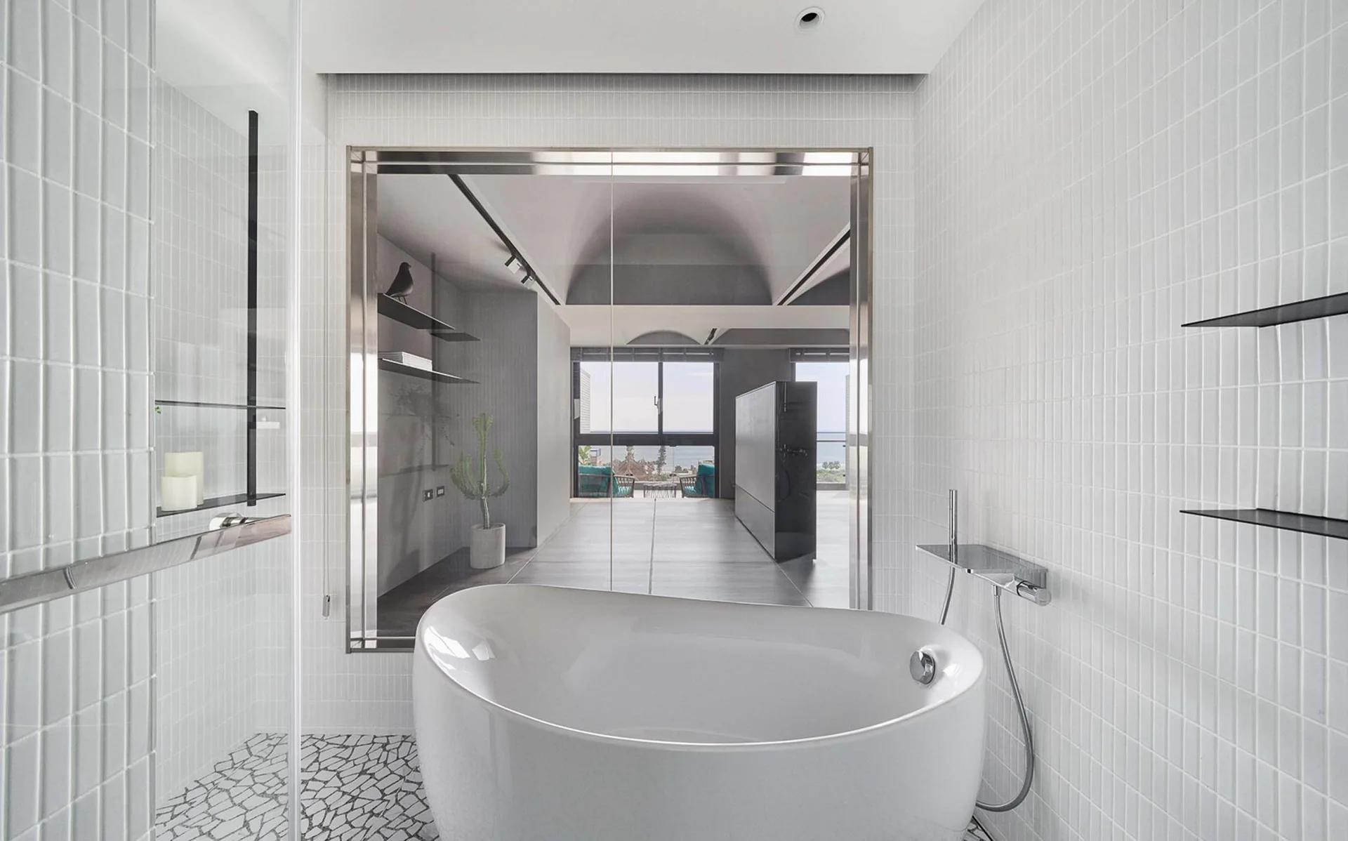 A modern white bathroom features a freestanding bathtub, small vertical wall tiles, mosaic stone flooring, and a large glass partition.