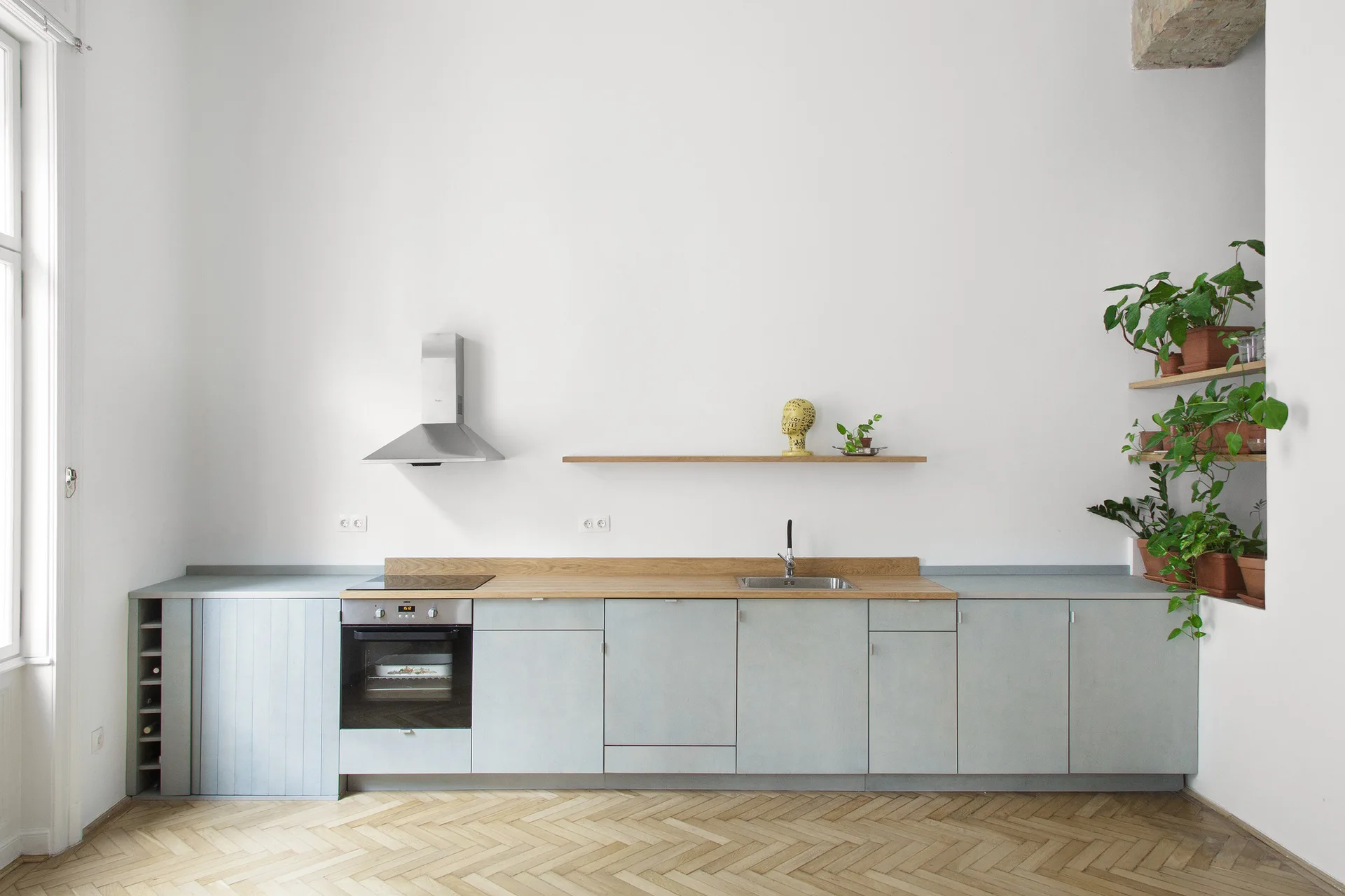 A minimalist kitchen features pale blue base cabinets, a light wood countertop, a stainless steel range hood, and light-toned herringbone wood flooring.