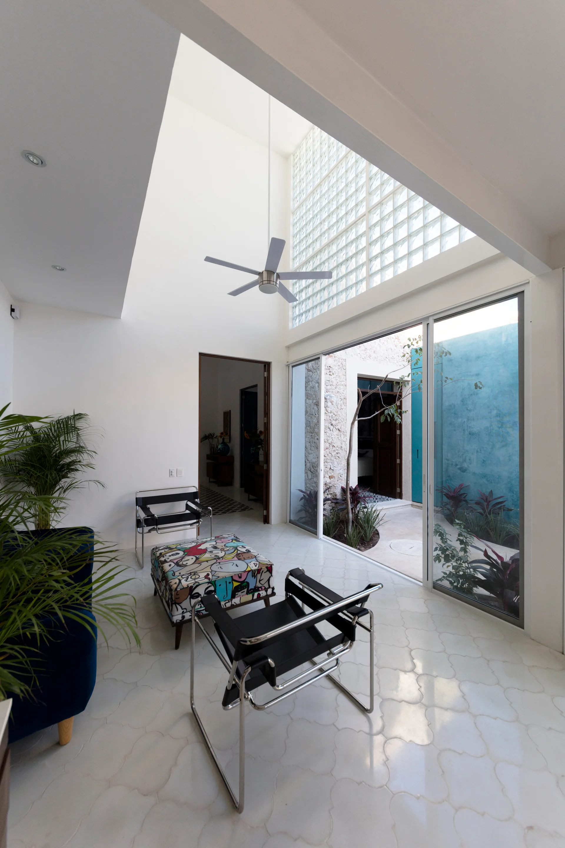 A modern living room features black leather and chrome chairs, a graphic-patterned ottoman, and large sliding glass doors opening to a courtyard.