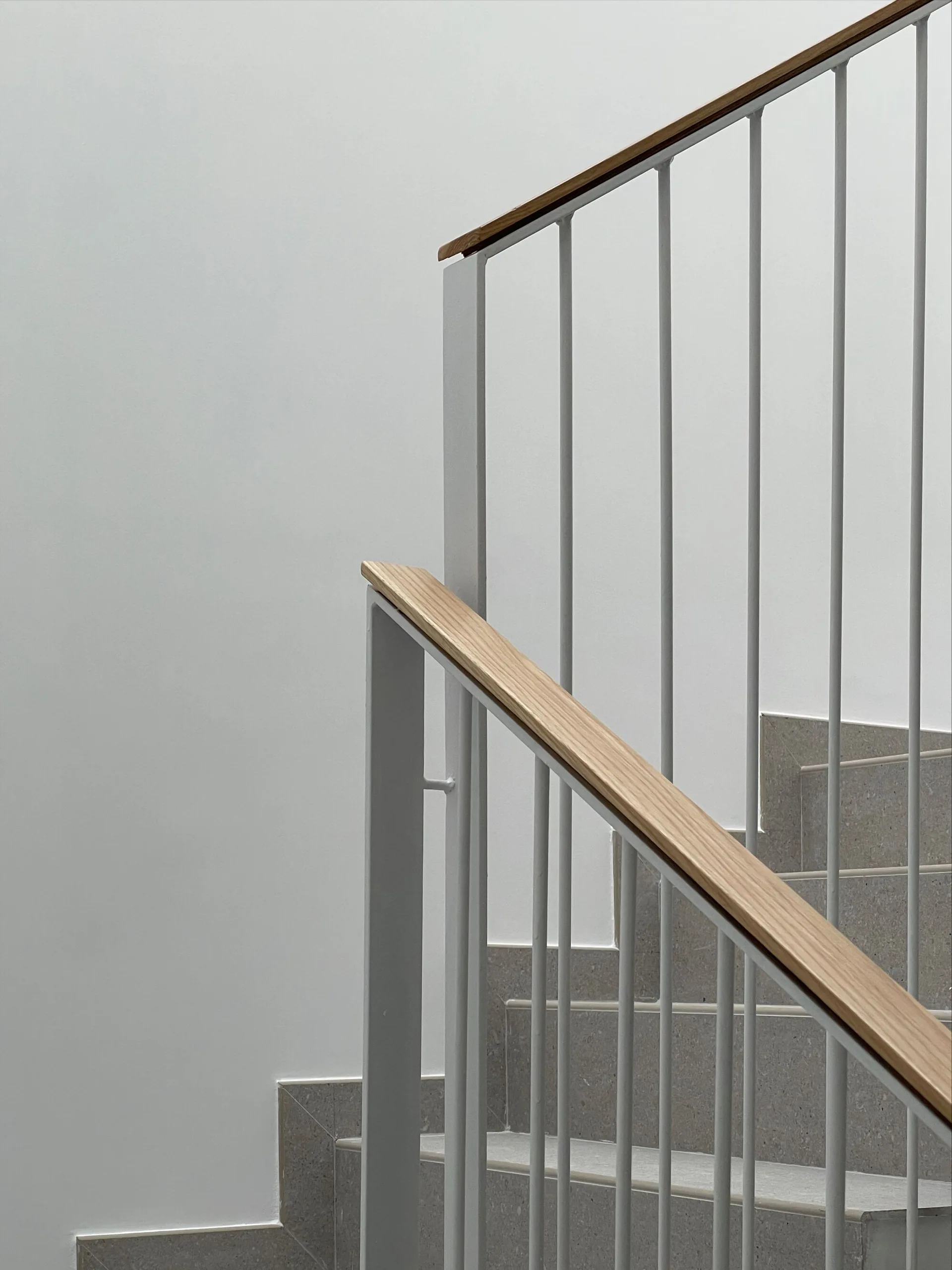 Minimalist staircase featuring grey stone steps, white vertical metal balusters, and a light wood handrail against a plain white wall.