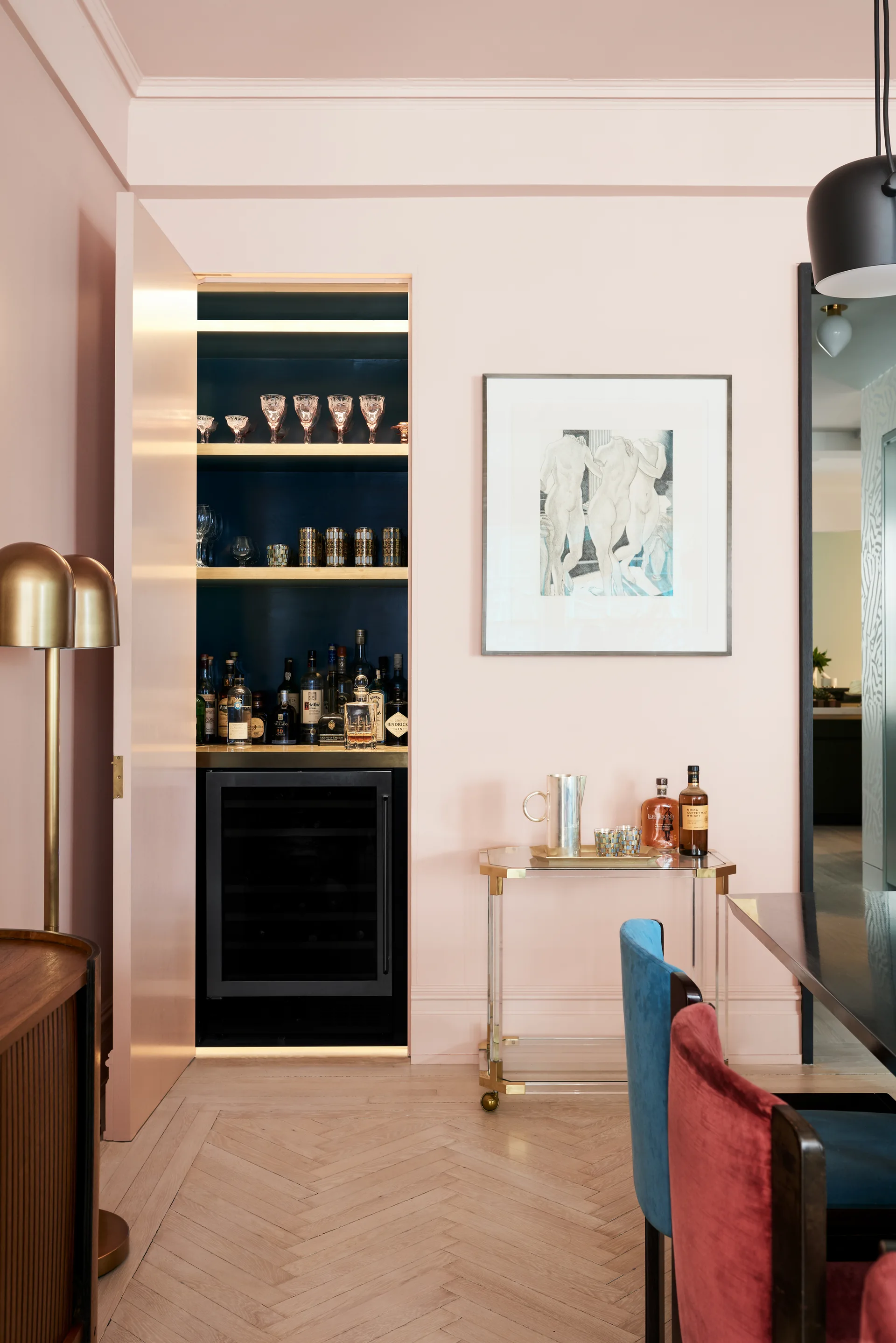 A blush pink dining room with herringbone wood floors and a built-in navy blue bar nook containing a wine fridge and lit glass shelving.