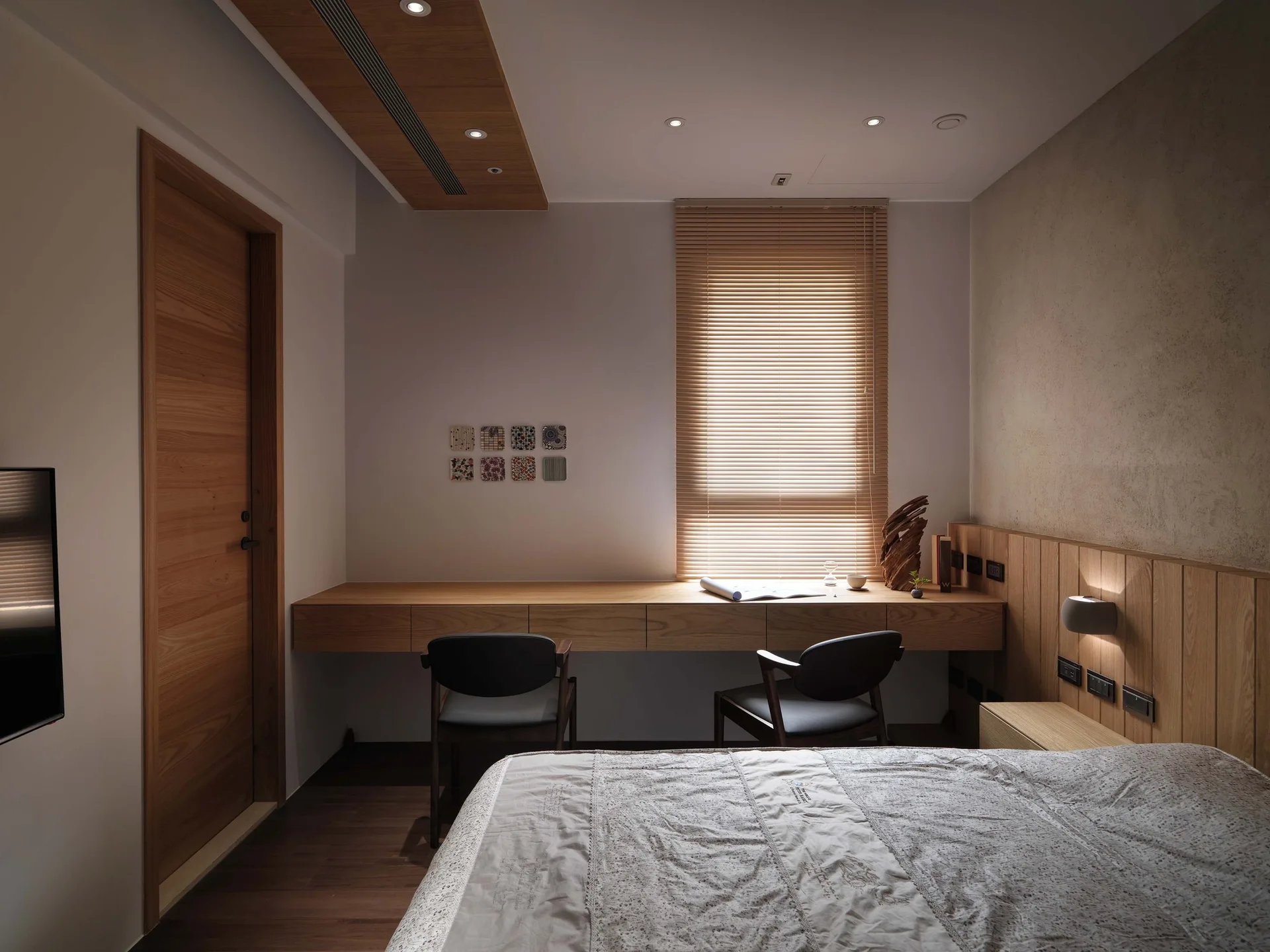 A modern minimalist bedroom featuring a long built-in wooden desk under a window with blinds, alongside vertical wood wall paneling.