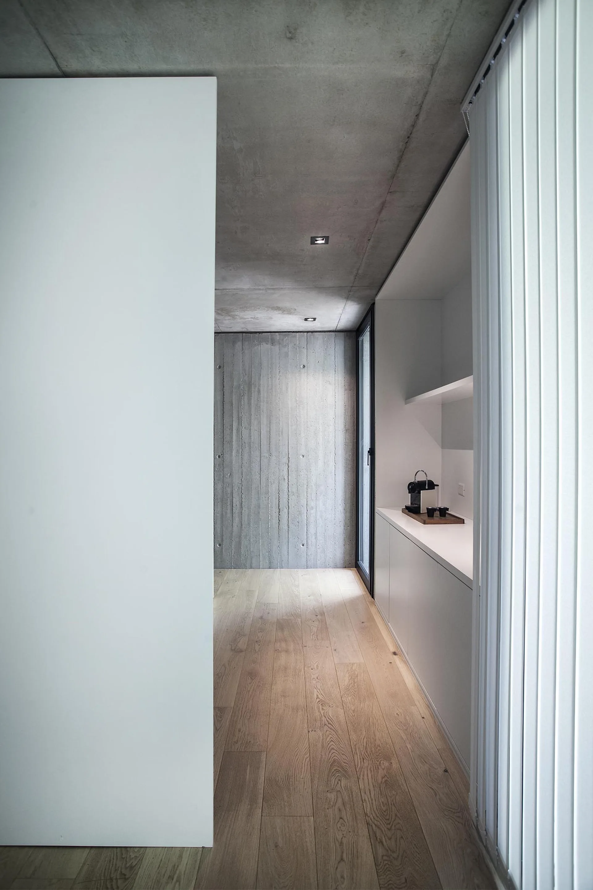 A minimalist corridor kitchenette features sleek white built-in cabinetry, light wood plank floors, and an exposed raw concrete ceiling and back wall.