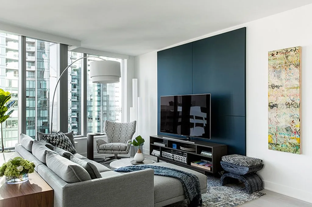 A contemporary living room featuring a gray sectional sofa, a dark blue paneled accent wall behind a TV, and floor-to-ceiling windows.