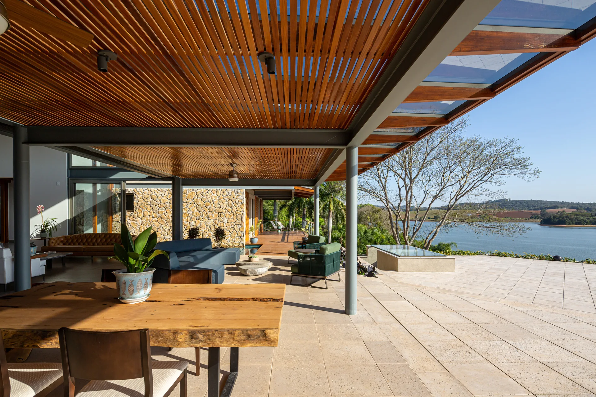 A covered outdoor patio featuring a slatted wood ceiling, a live-edge dining table, stone walls, and scenic lake views.