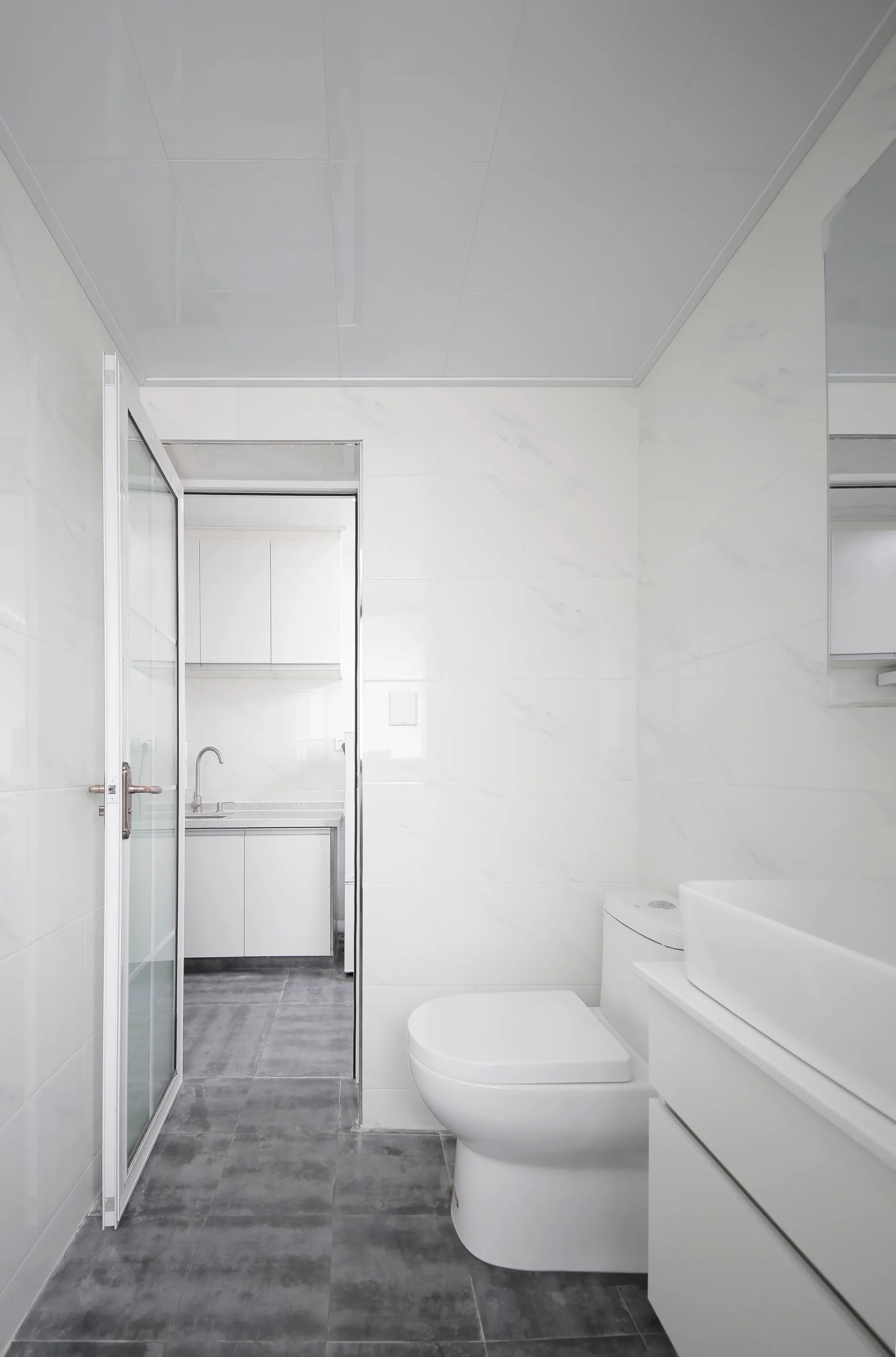 A modern bathroom featuring glossy white marble-patterned wall tiles, a white toilet and vanity sink, and dark grey floor tiles.