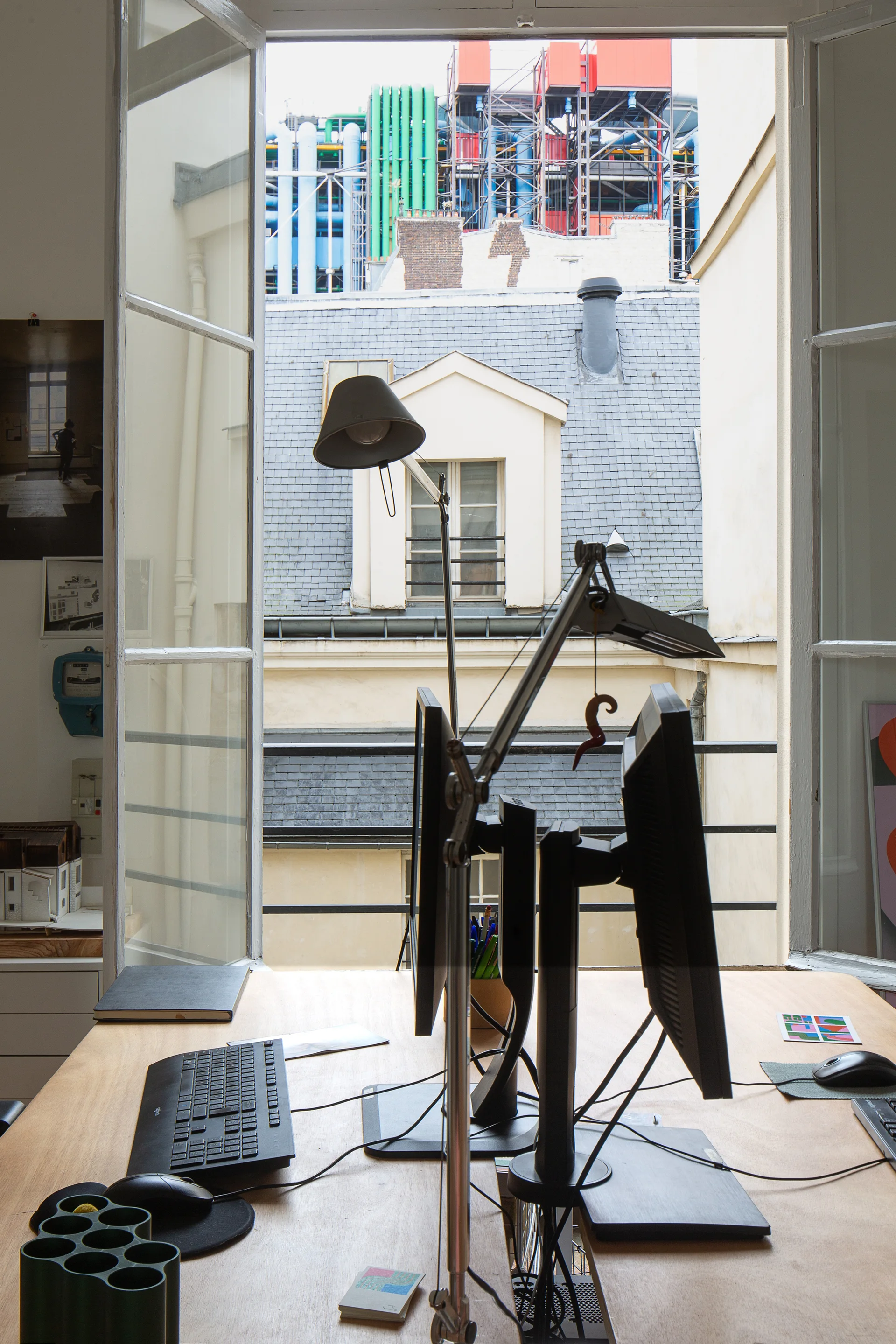 A light wood desk with computer monitors and keyboards sits in front of an open casement window displaying classic city rooftops.