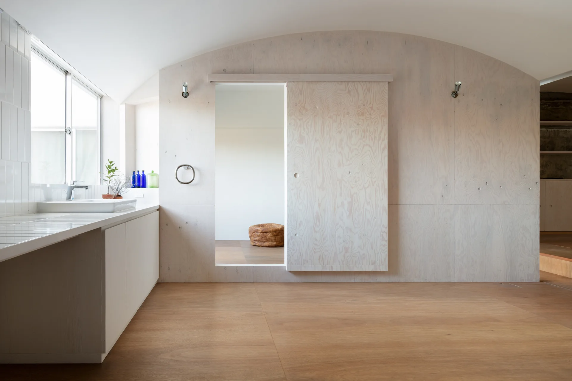 A minimalist kitchen features a white tiled countertop with a sink, a large plywood paneled wall with a sliding door, and light wood flooring.
