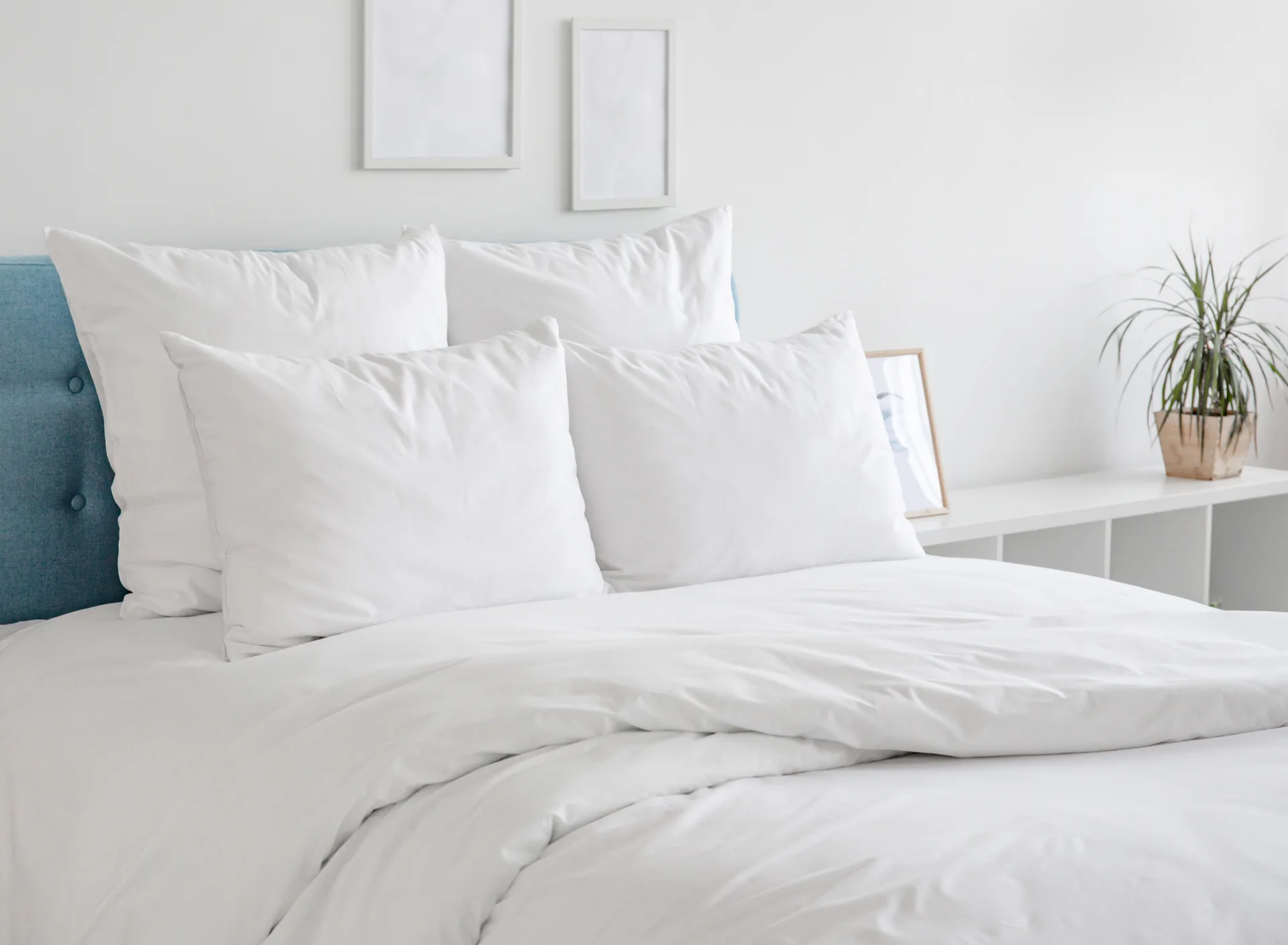 A minimalist bed with a blue upholstered headboard and white linens sits against a white wall decorated with empty frames.