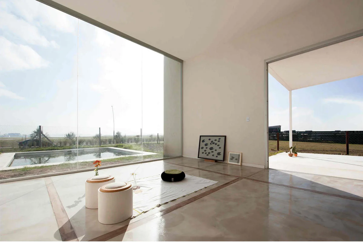 A minimalist interior space with polished concrete flooring, white walls, two small stools on a floor mat, and an expansive glass wall overlooking a pool.