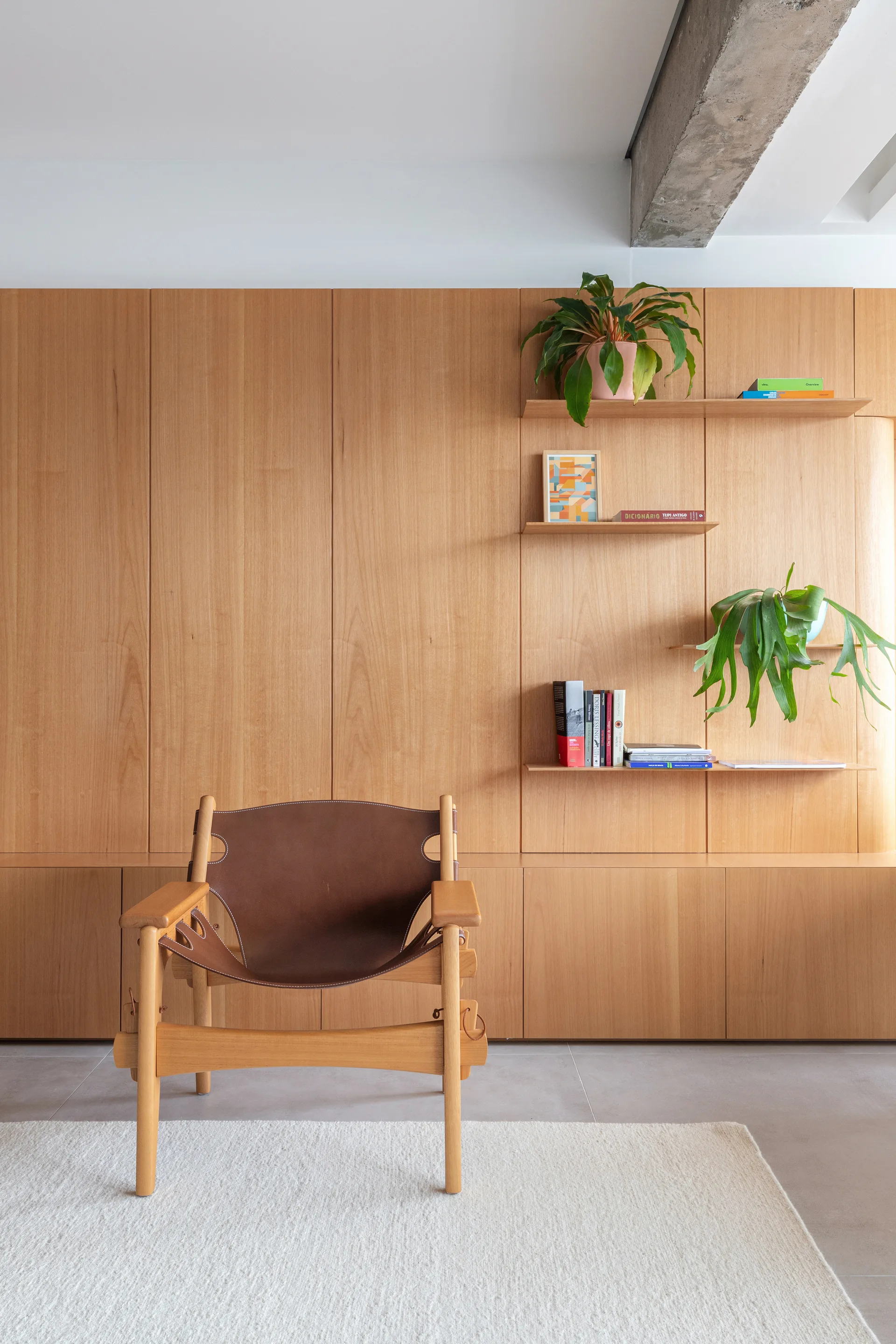 A mid-century leather lounge chair sits on a textured white rug in front of a full-height natural wood storage wall with built-in shelves.