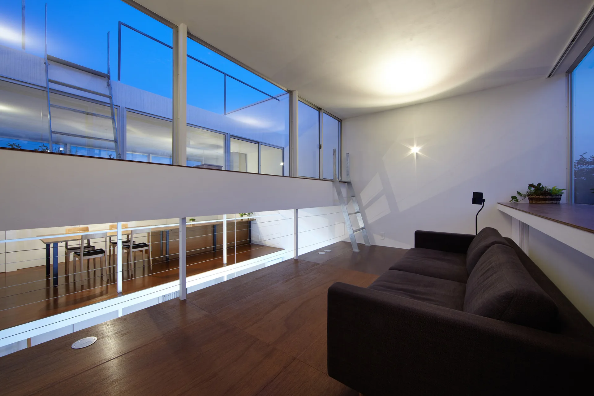 A modern mezzanine living space with a dark brown sofa, wood flooring, and large glass windows overlooking a lower dining room.