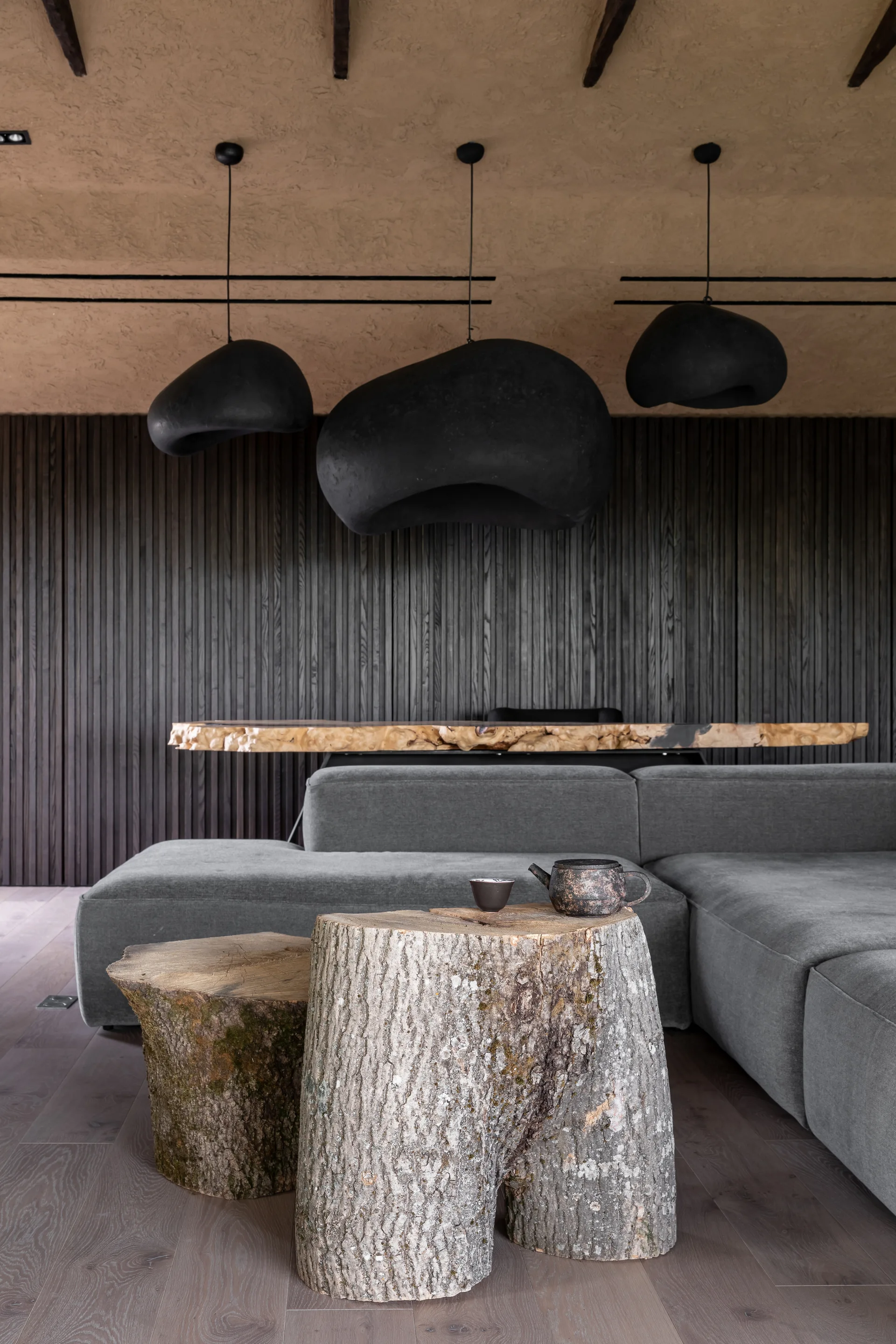 A grey sectional sofa is paired with tree stump coffee tables under black organic pendant lights in a room with a dark slatted wood wall.