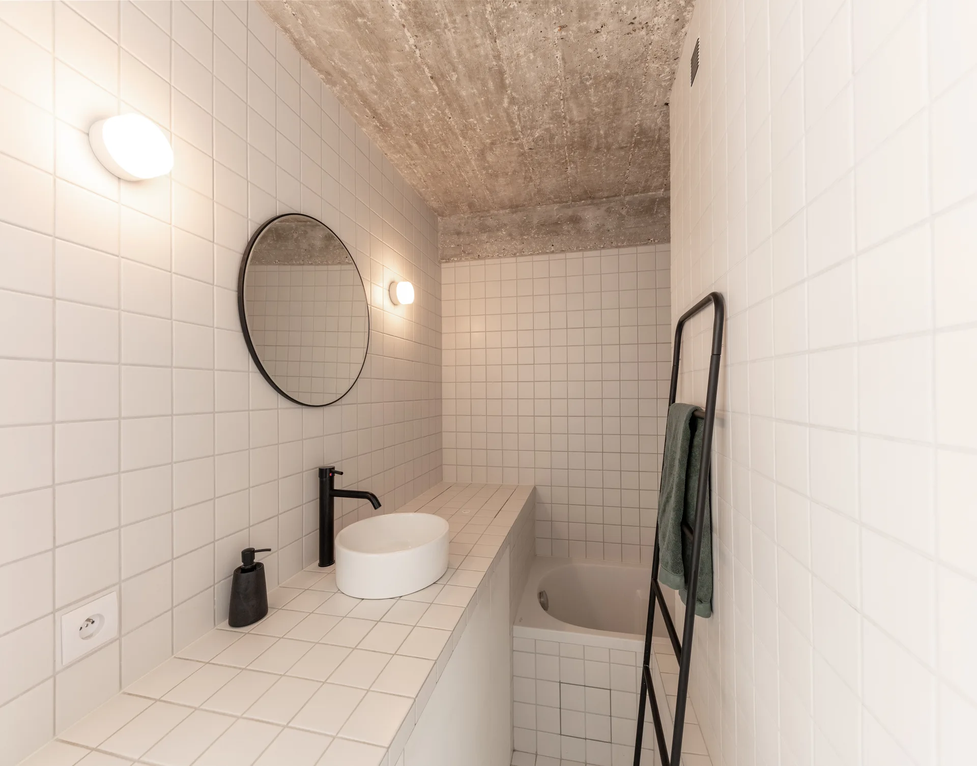 A narrow modern bathroom with white square tiled walls, a round mirror over a vessel sink, a leaning black towel ladder, and an exposed concrete ceiling.