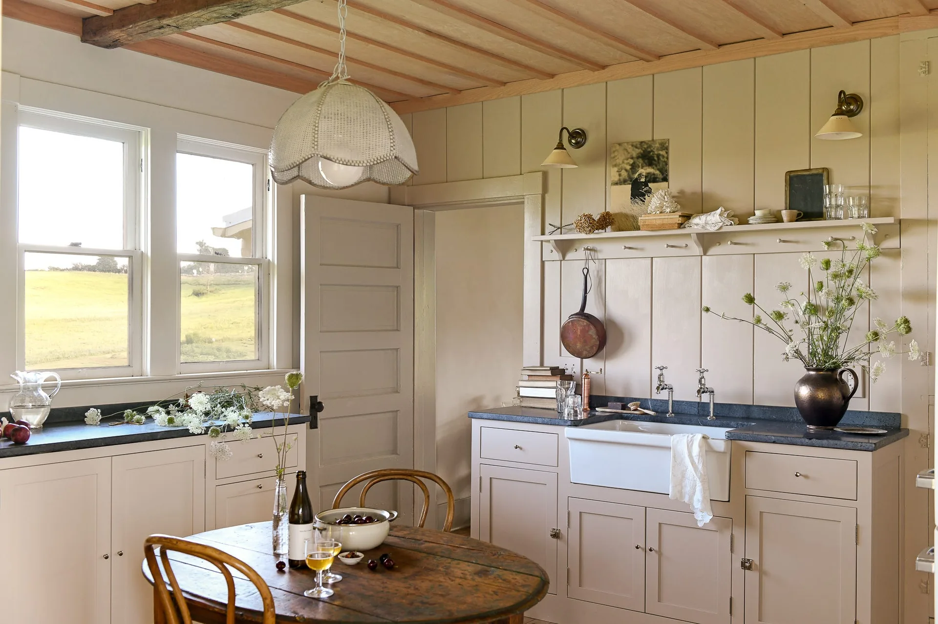 A rustic kitchen features warm taupe paneled walls, a white farmhouse sink, dark countertops, and a wooden dining table beneath a wood-planked ceiling.