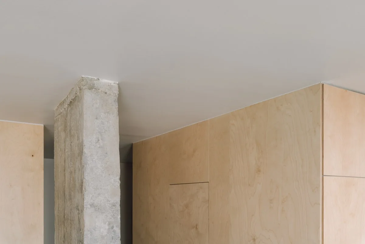 An unfinished gray concrete column stands next to light wood plywood wall panels under a flat white ceiling.