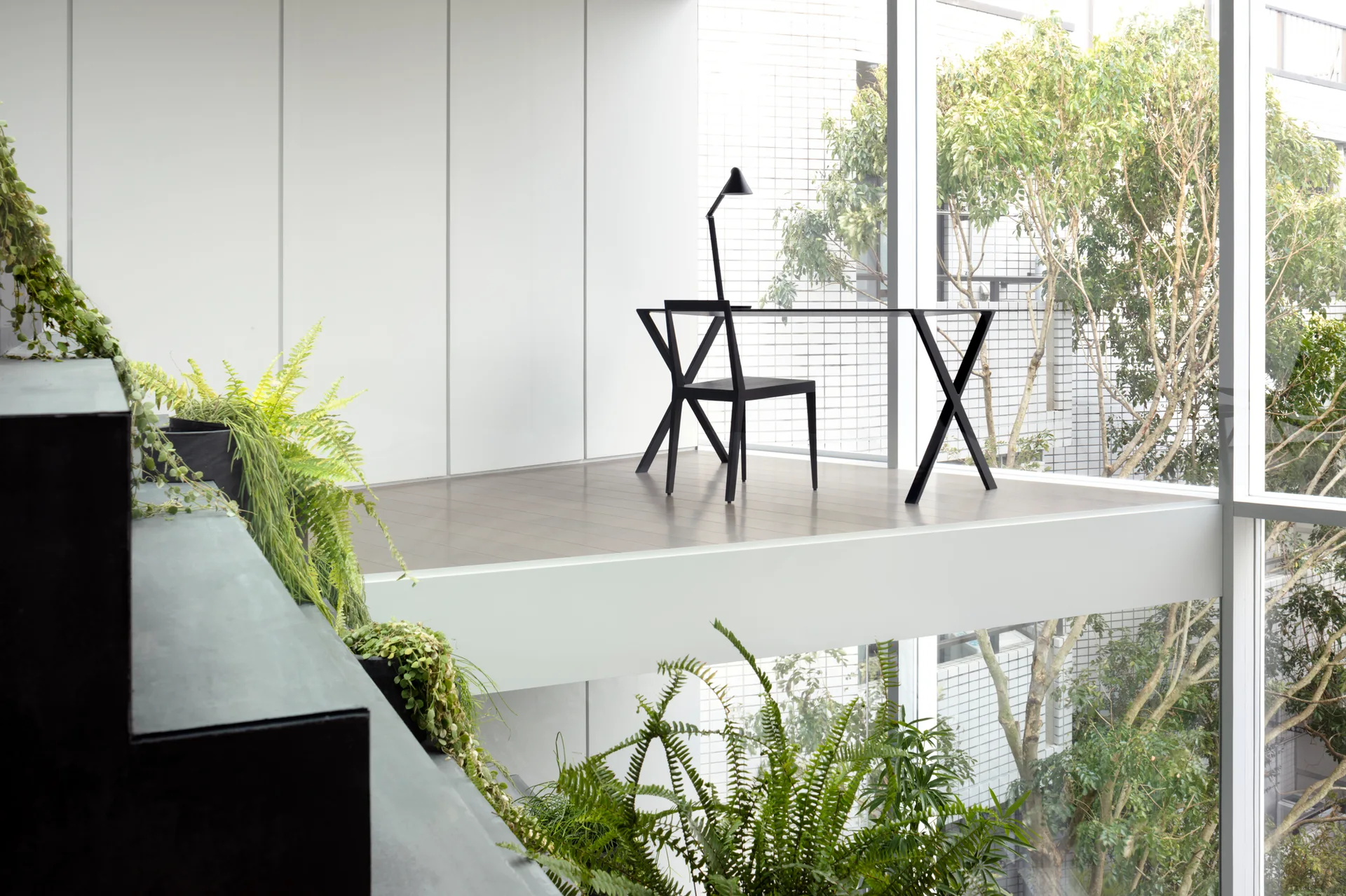 A minimalist black desk and chair sit on a light wood mezzanine landing next to a floor-to-ceiling glass window overlooking outdoor trees.