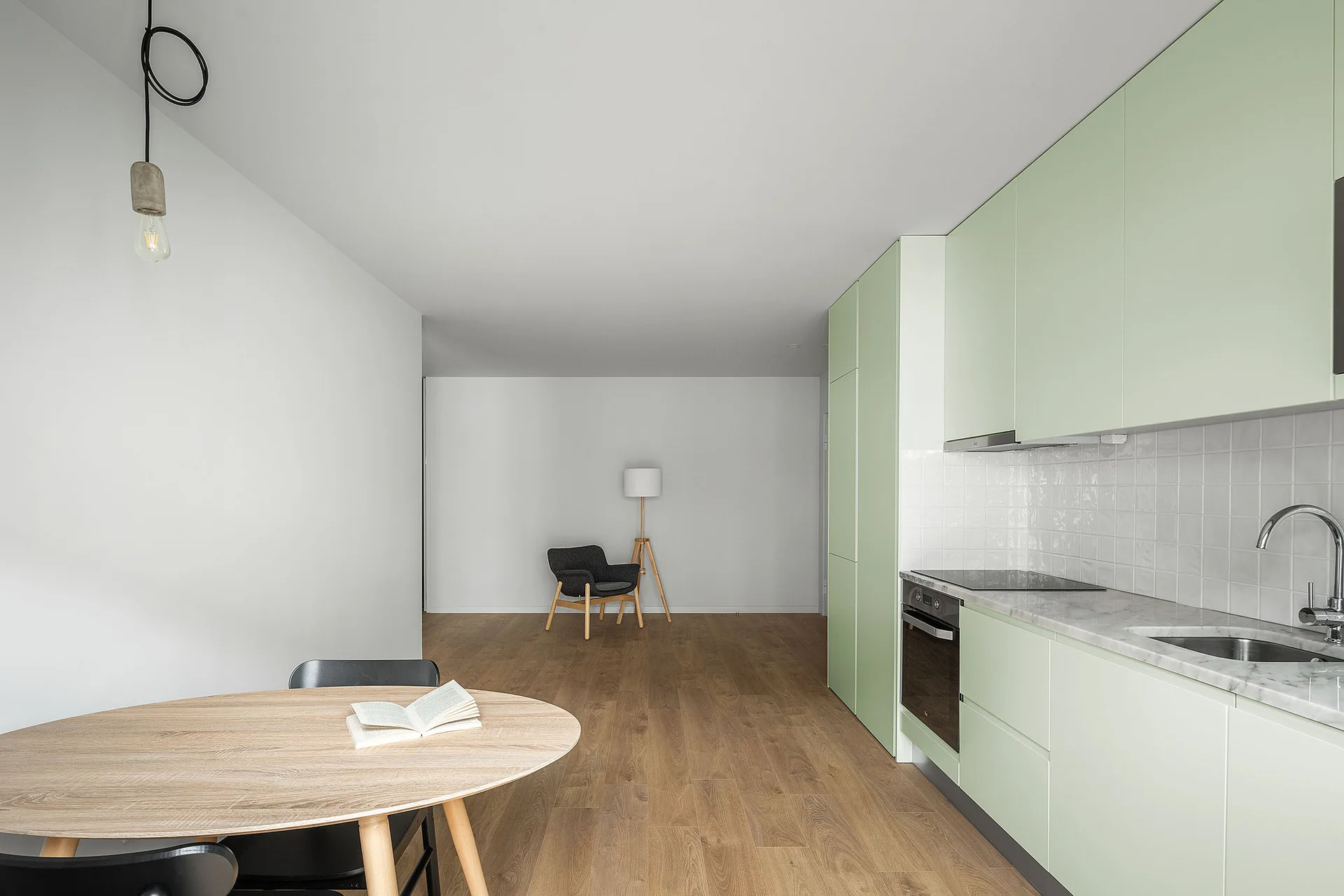 A minimalist kitchen features mint green flat-panel cabinetry, a light wood dining table, white square tiled backsplash, and wood plank flooring.