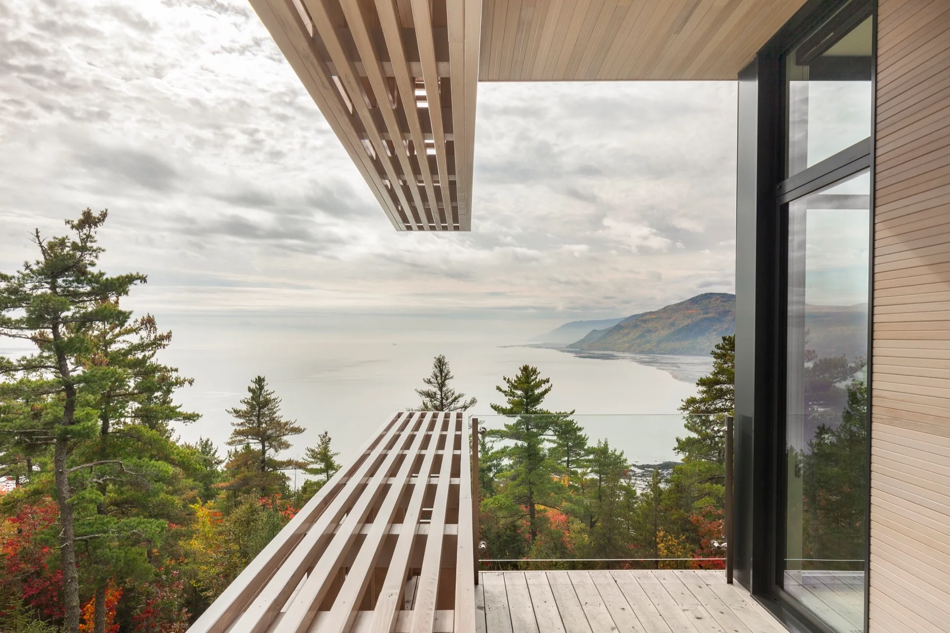 A modern wooden balcony featuring a built-in slatted bench, matching overhead canopy, and glass railing overlooking a cloudy coastal landscape.