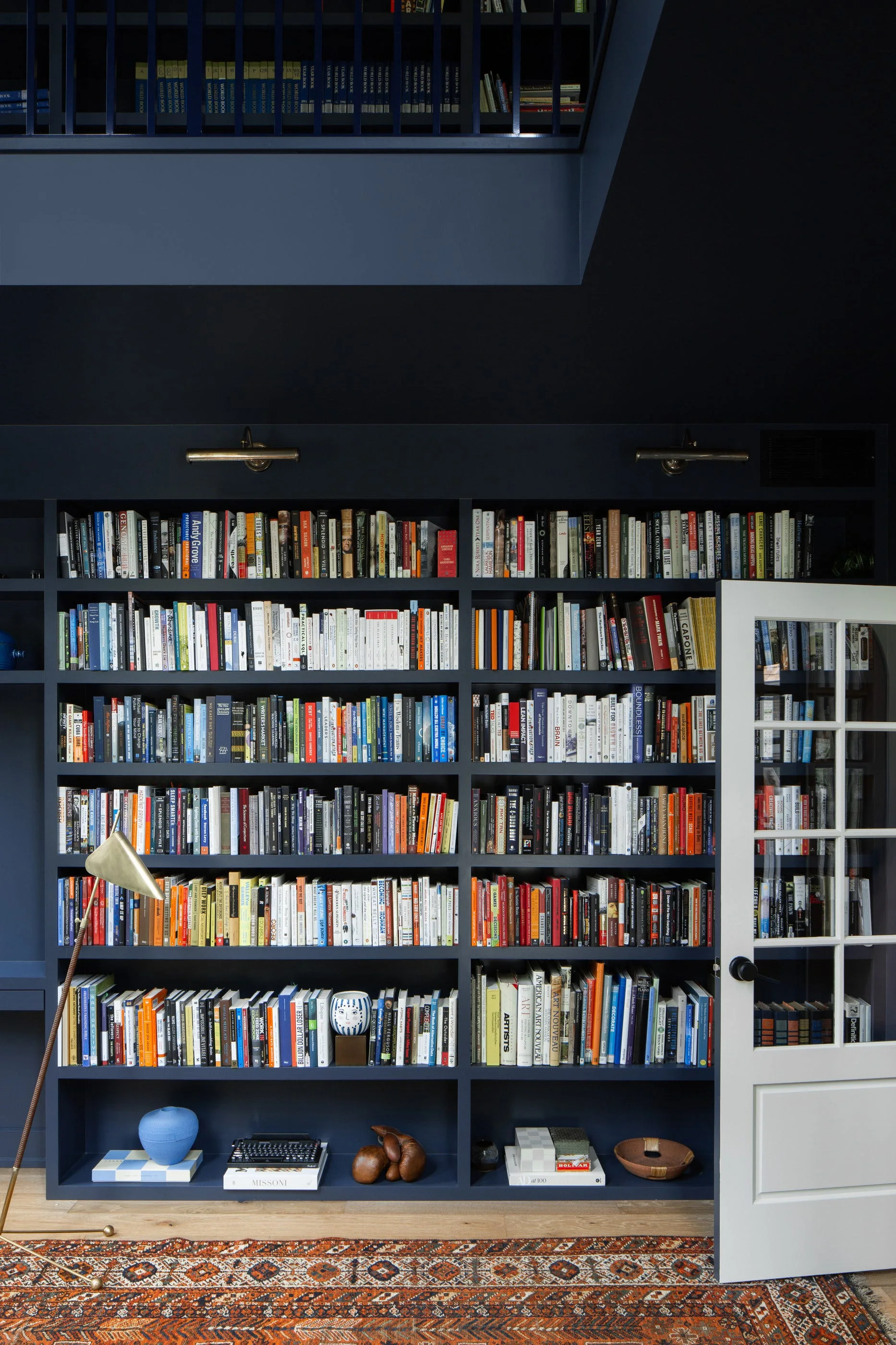 A floor-to-ceiling navy blue built-in bookcase filled with colorful books, flanked by a white French door and a patterned orange area rug.