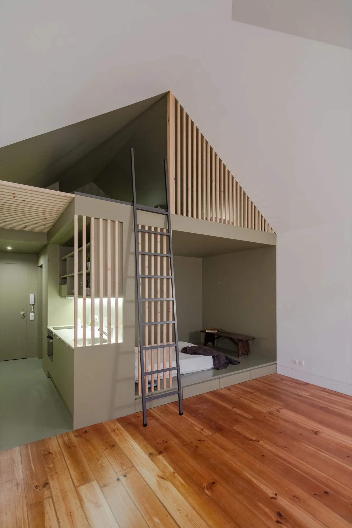 A compact studio features an olive green built-in structure with an integrated kitchenette, a platform sleeping nook, and a wood-slatted loft.