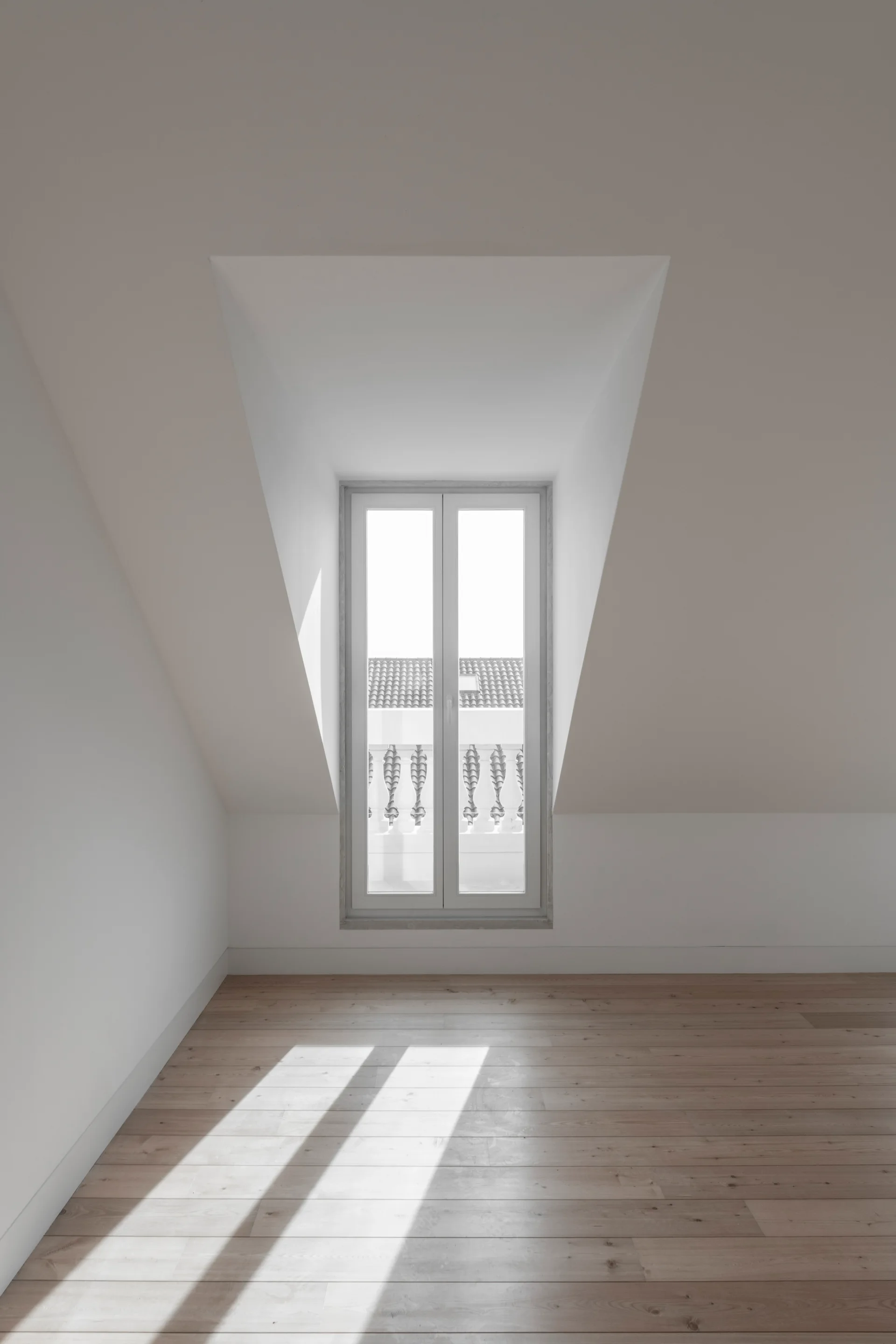 An empty minimalist attic room featuring white sloped walls, a central dormer window letting in natural light, and light wood plank flooring.