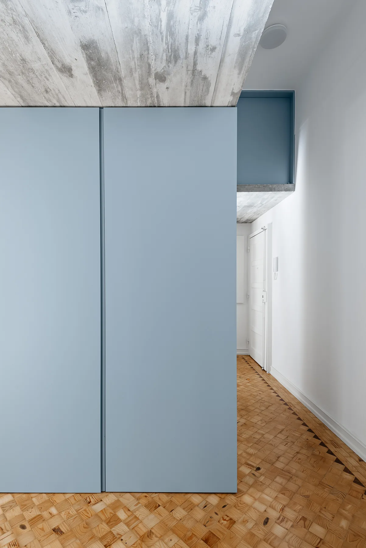 A large matte blue built-in wardrobe stands in a hallway featuring an exposed concrete ceiling and detailed wooden parquet flooring.