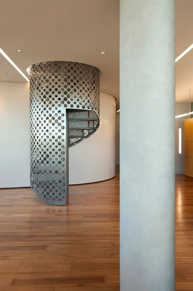 A modern interior featuring a perforated metal spiral staircase, warm hardwood flooring, a curved white wall, and a large concrete column in the foreground.