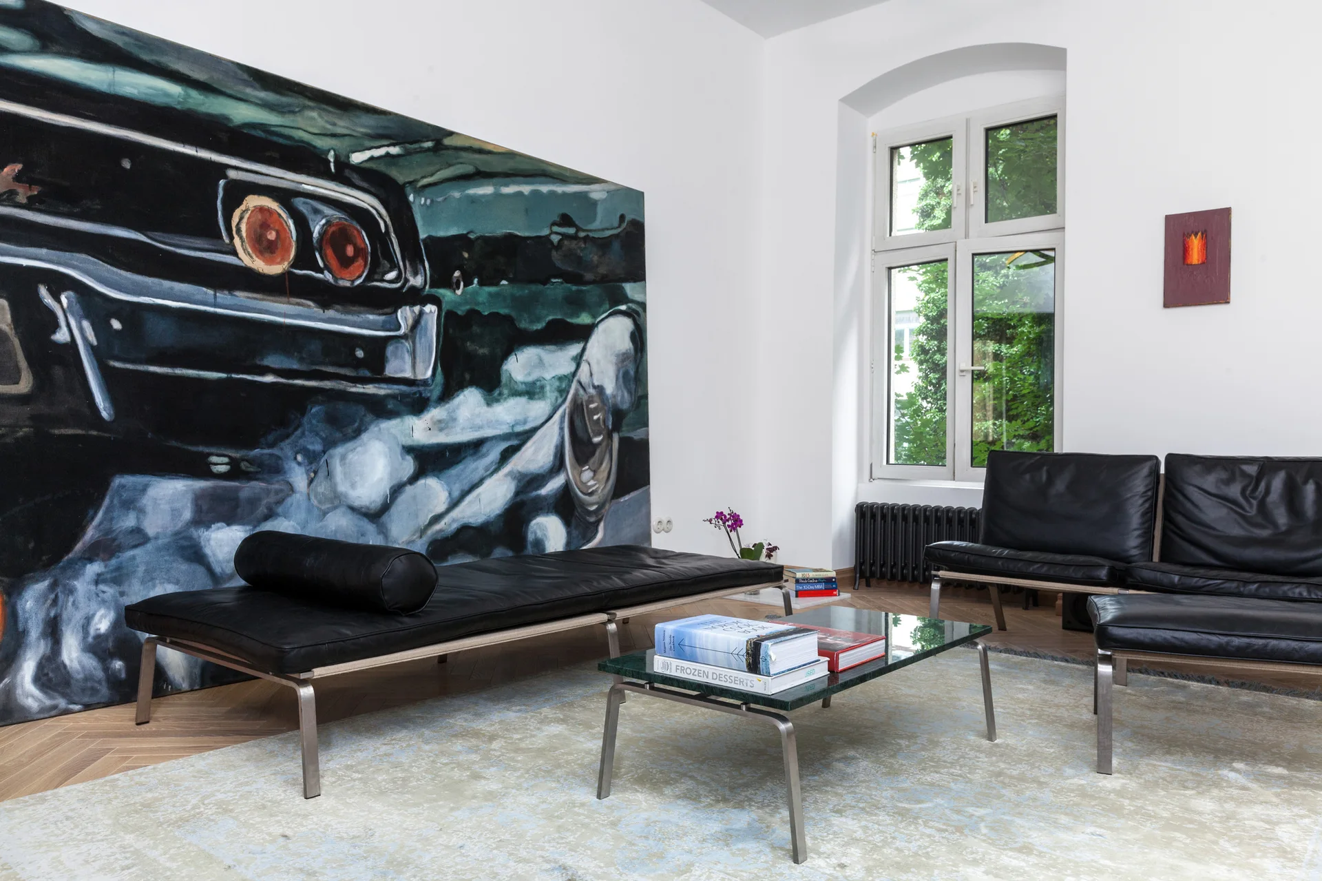 A modernist living room featuring black leather seating, a low coffee table, and an oversized painting of a car tail on white walls.