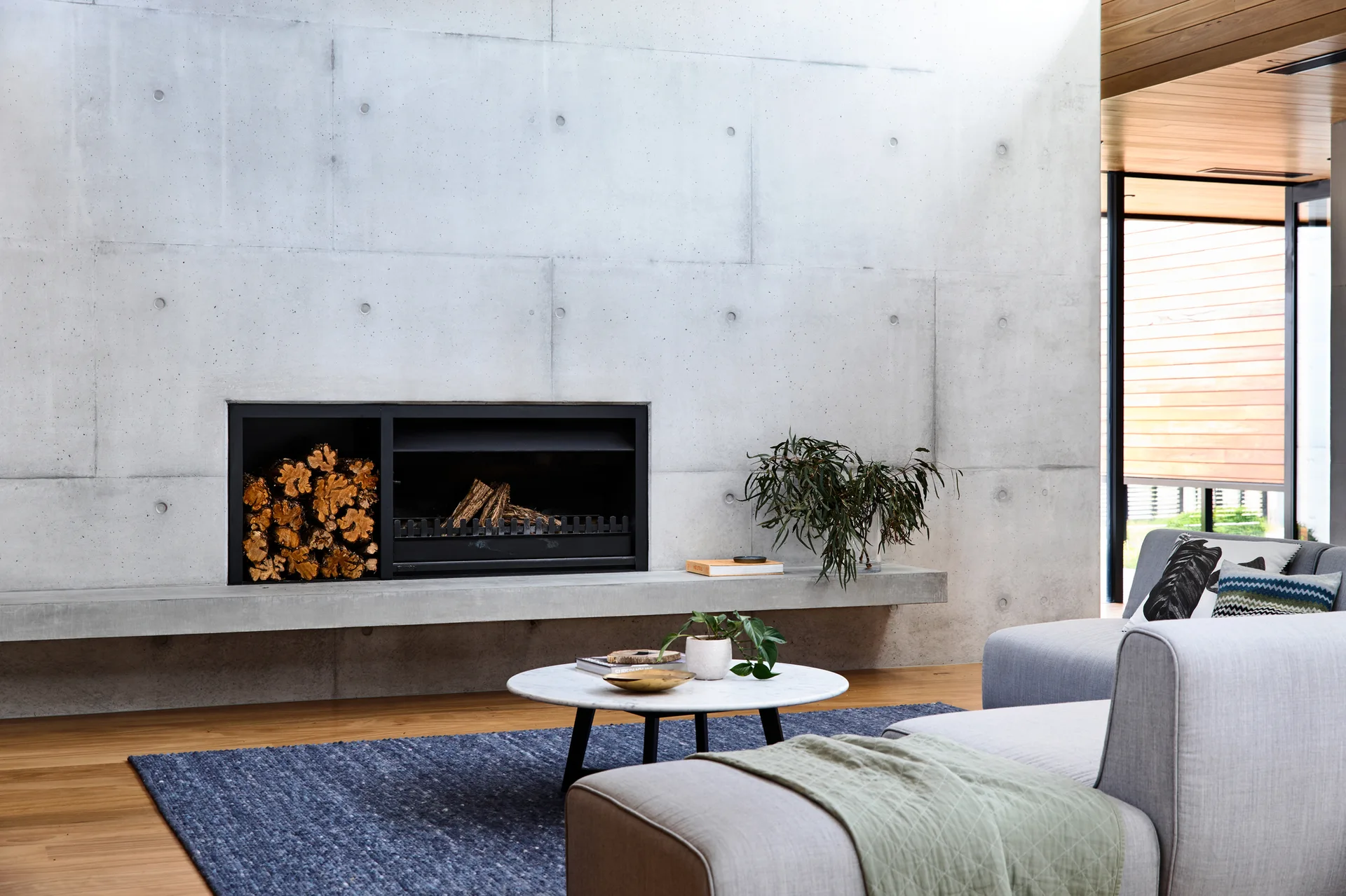A minimalist living room featuring a concrete fireplace wall with a floating hearth, a grey sectional sofa, and a textured blue rug.