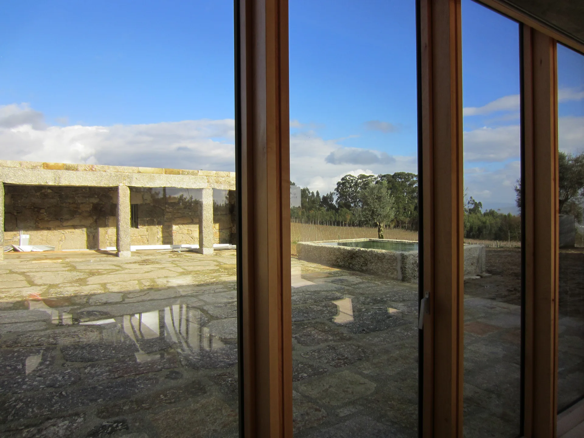 A view through large wood-framed glass windows onto a rustic stone-paved patio with a stone structure and a small water basin under a blue sky.