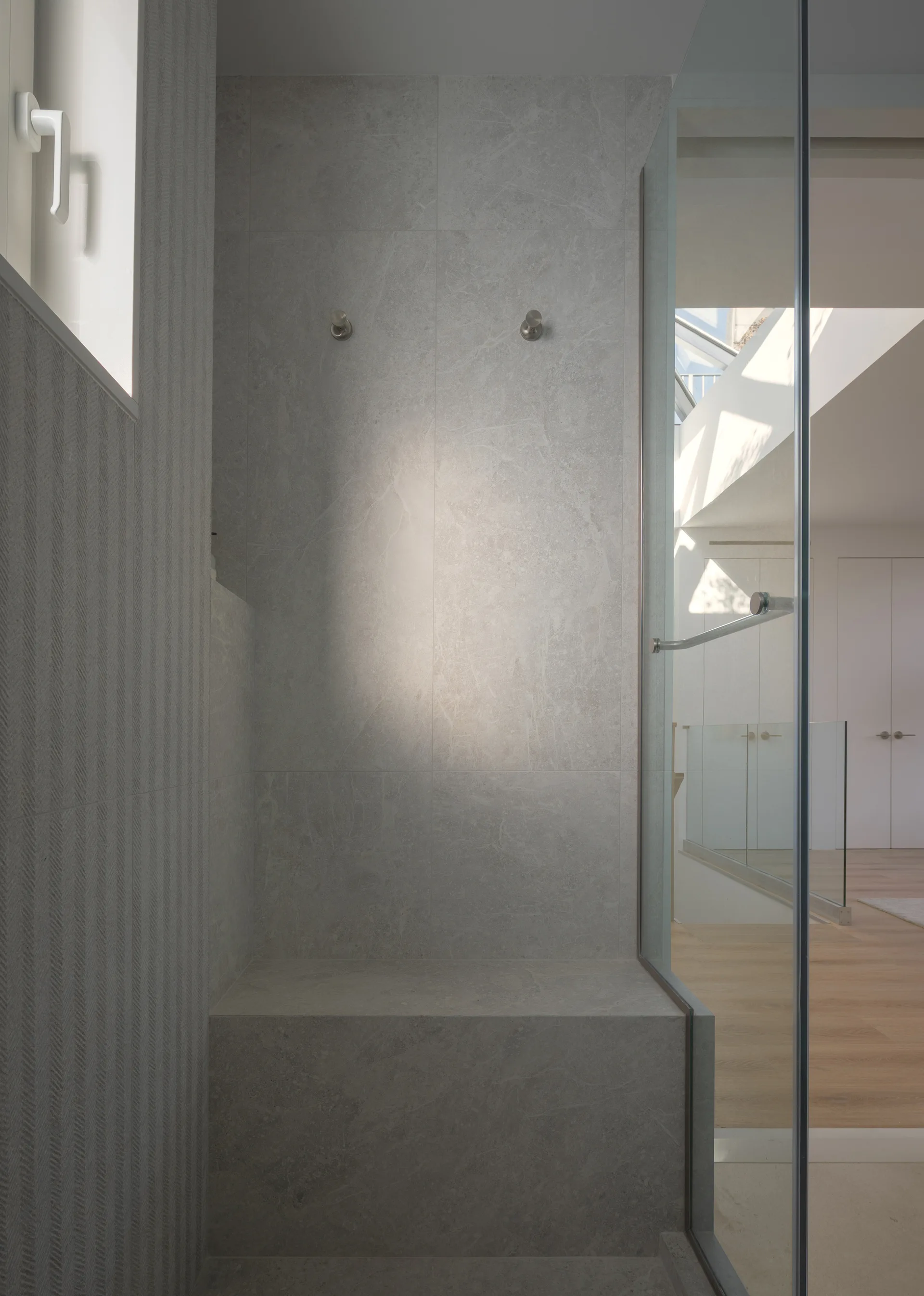 A walk-in shower featuring large-format grey stone tiles, a built-in bench, two silver hooks, and a glass partition door.