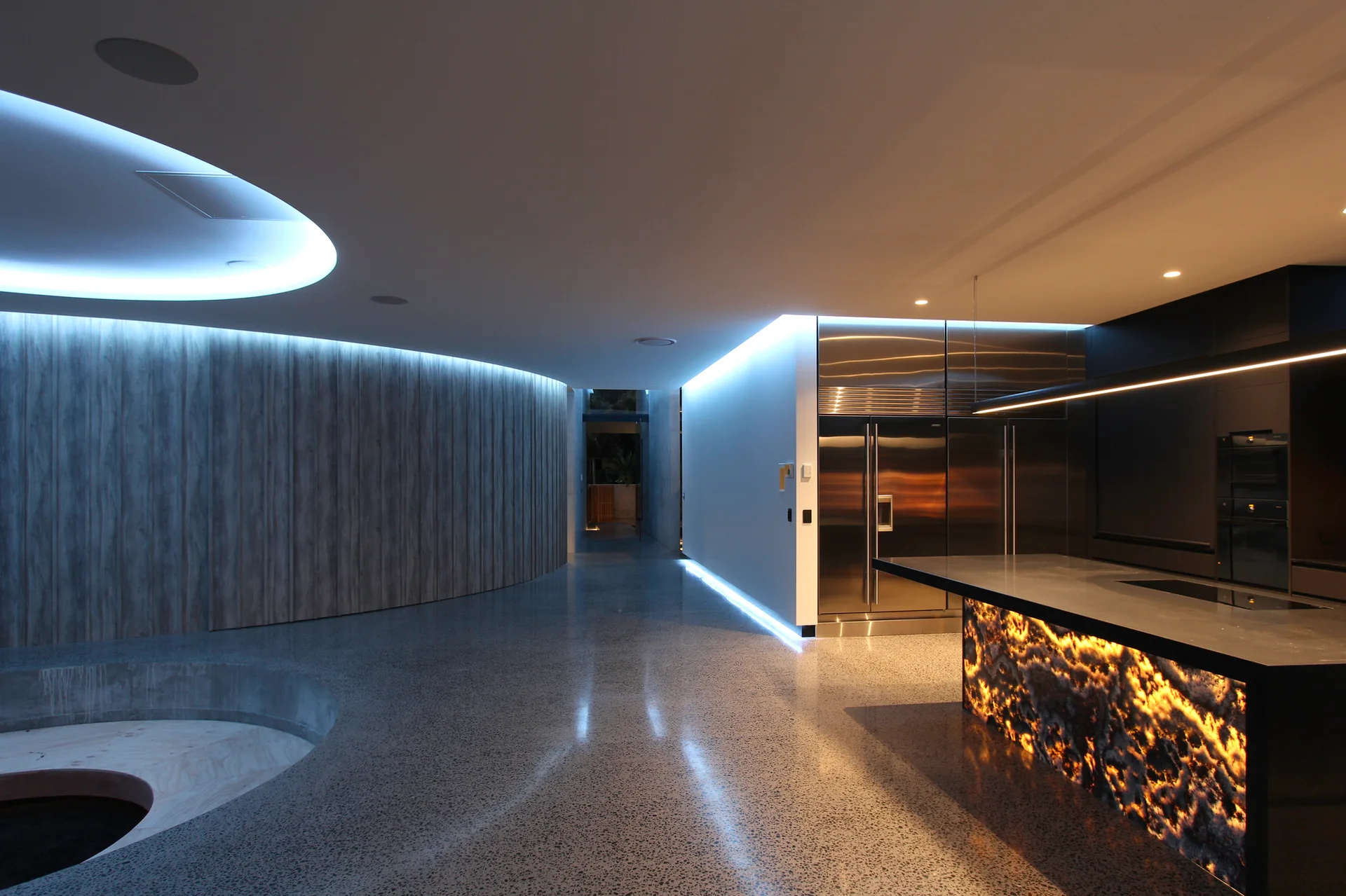 A modern minimalist kitchen featuring an illuminated stone island, blue LED cove lighting, a curved wood wall, and polished concrete flooring.