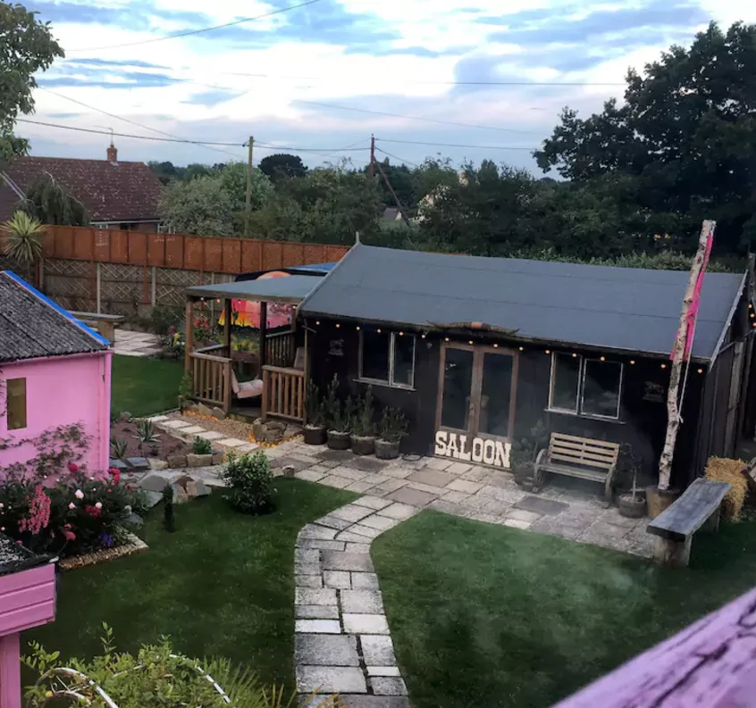 A backyard garden featuring a dark brown wooden saloon cabin with string lights, a pink shed, and a stone path winding through the lawn.