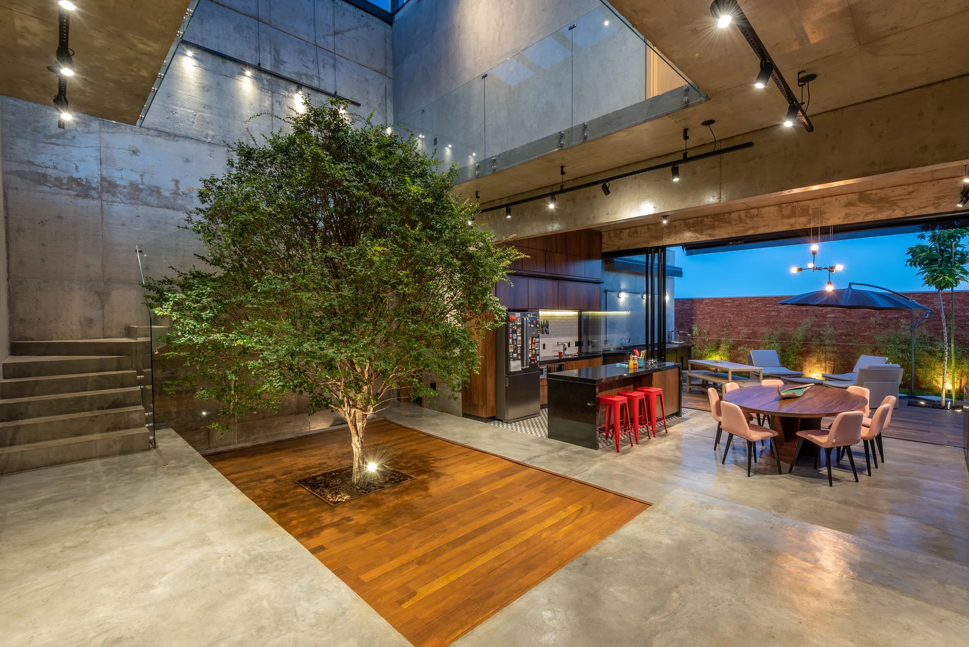A modern open-plan interior with exposed concrete walls, a large indoor tree in a wood floor inlay, and a connected kitchen.