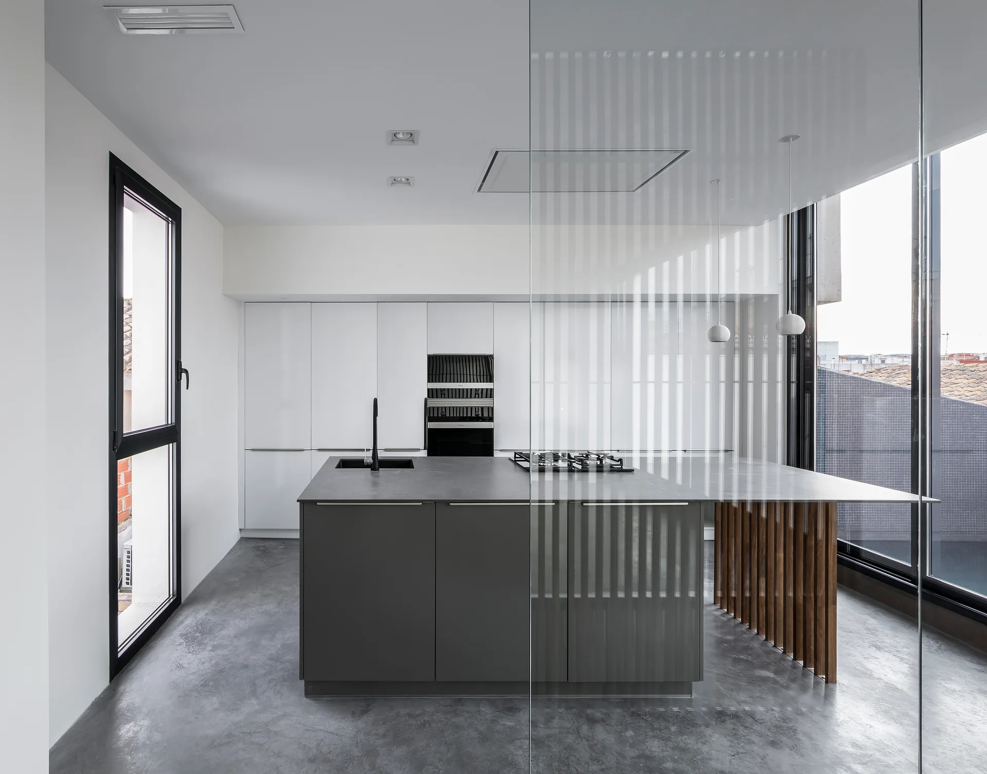 A modern kitchen features a dark gray island with vertical wood slats, white handleless cabinets, and polished concrete flooring.