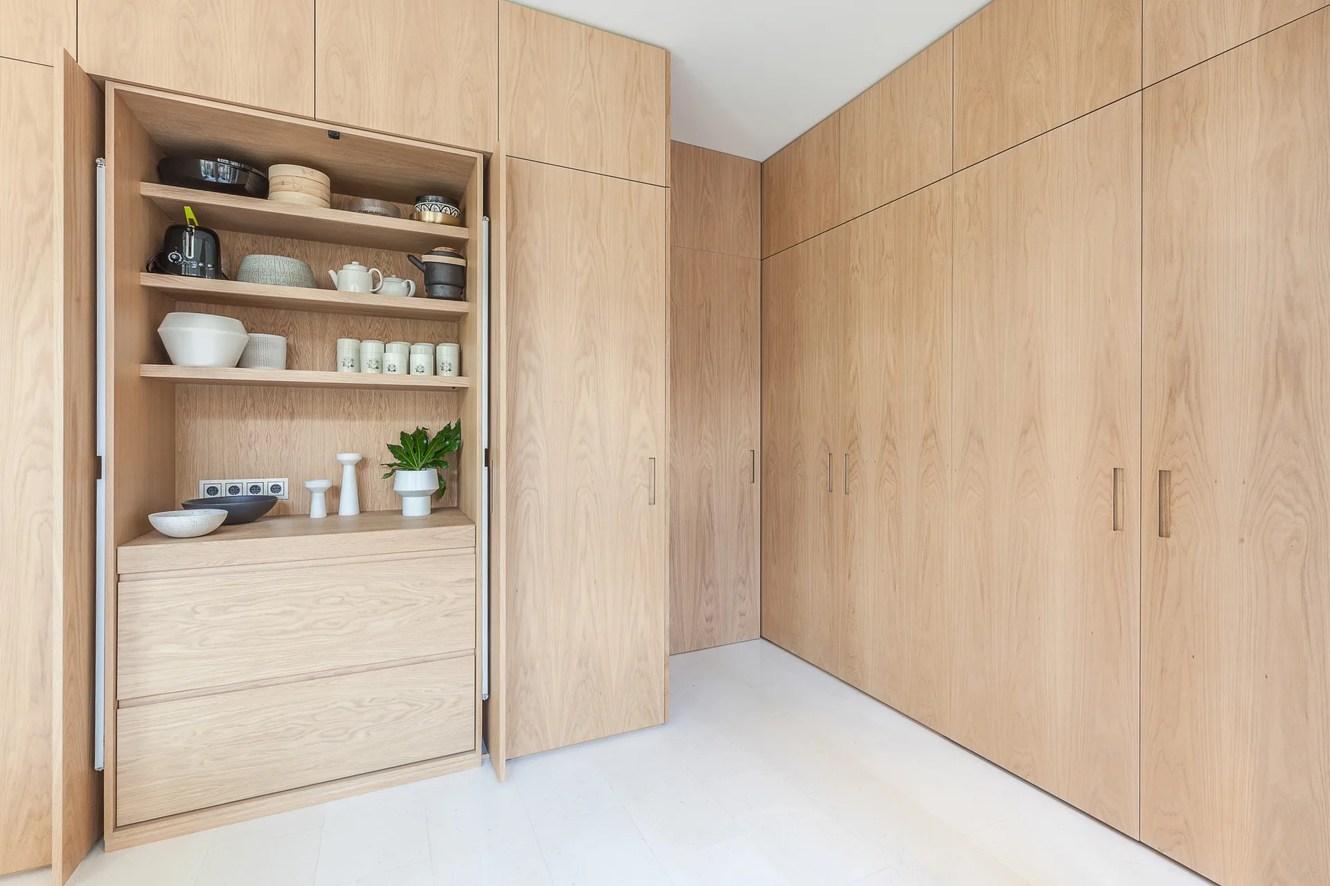 Minimalist kitchen featuring floor-to-ceiling light oak wood cabinetry with a recessed cabinet open to reveal shelves of ceramic kitchenware.