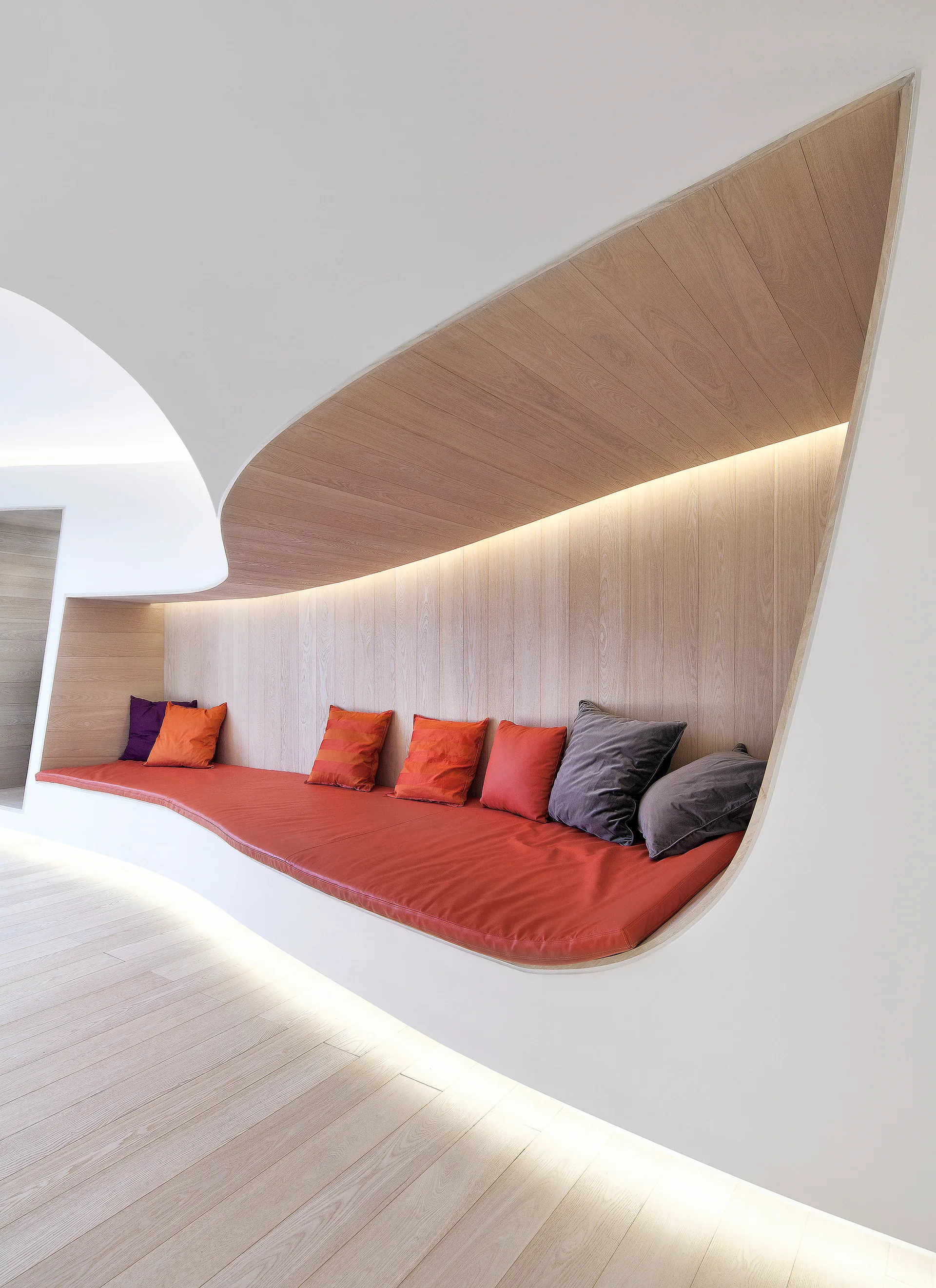 A custom curved wooden bench built into a white wall features a long red cushion, colorful pillows, and warm integrated cove lighting.