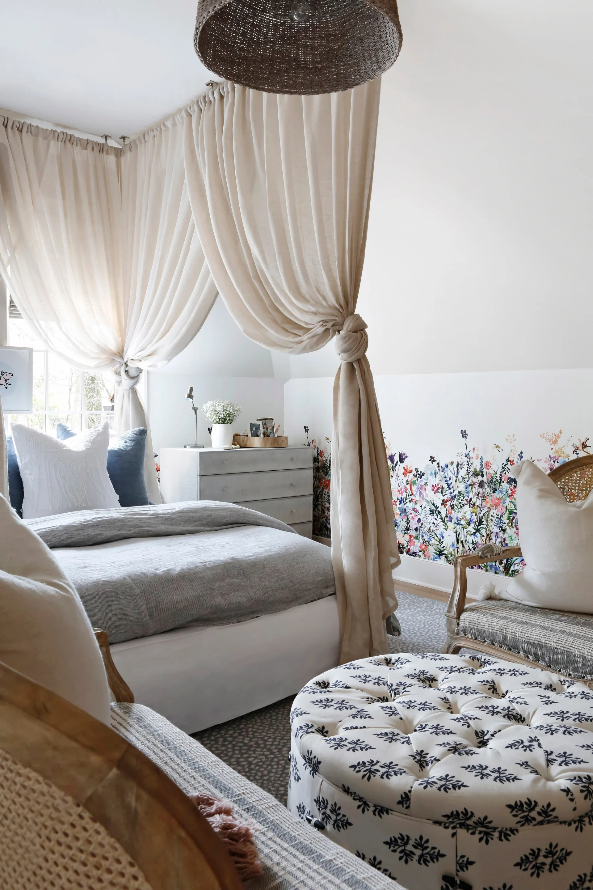 A bedroom features a bed enclosed by draped sheer beige canopy curtains, alongside a floral wallpaper border, grey bedding, and a tufted patterned ottoman.
