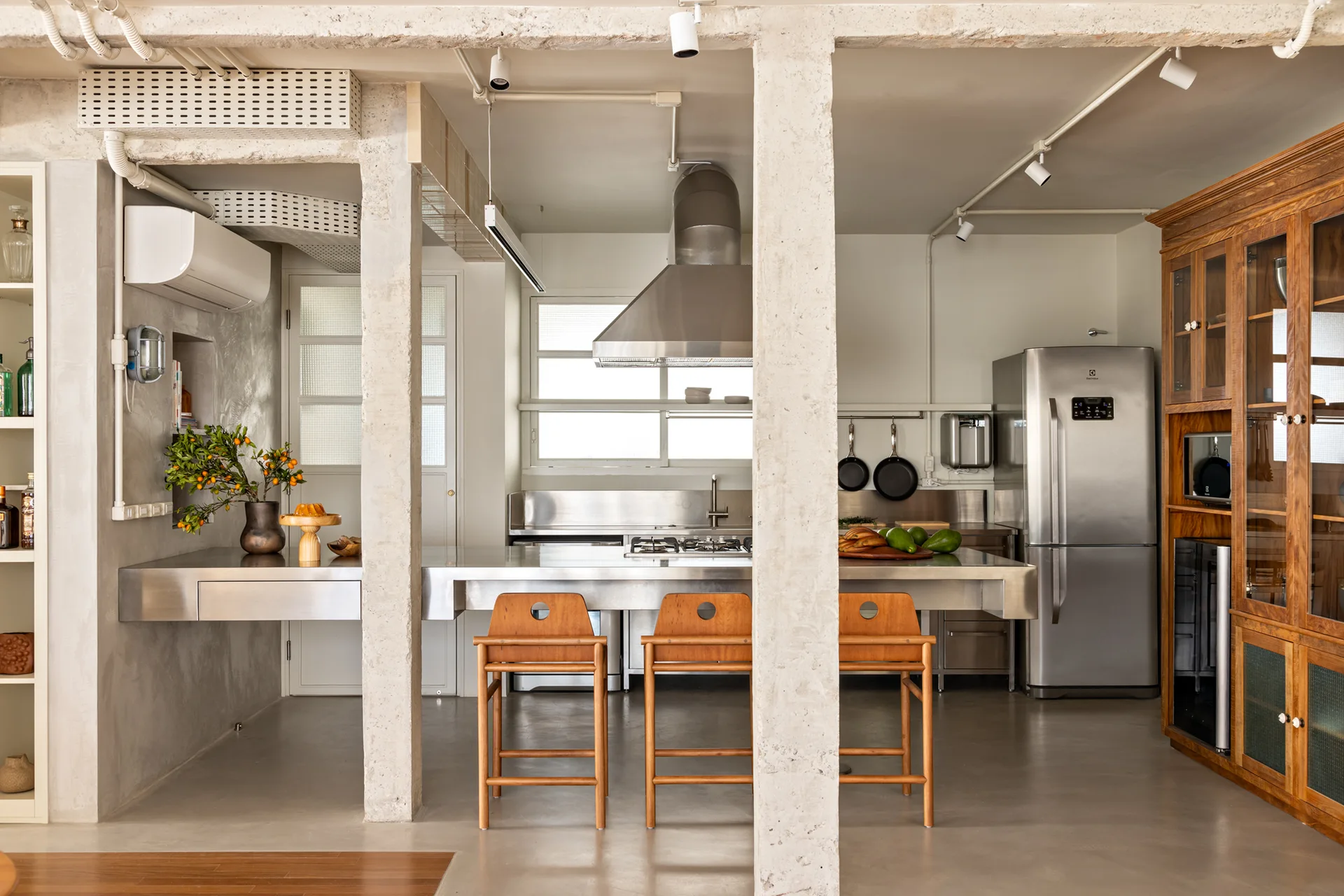 A modern industrial kitchen features a stainless steel island, raw concrete columns, three wood bar stools, and a large wood cabinet.