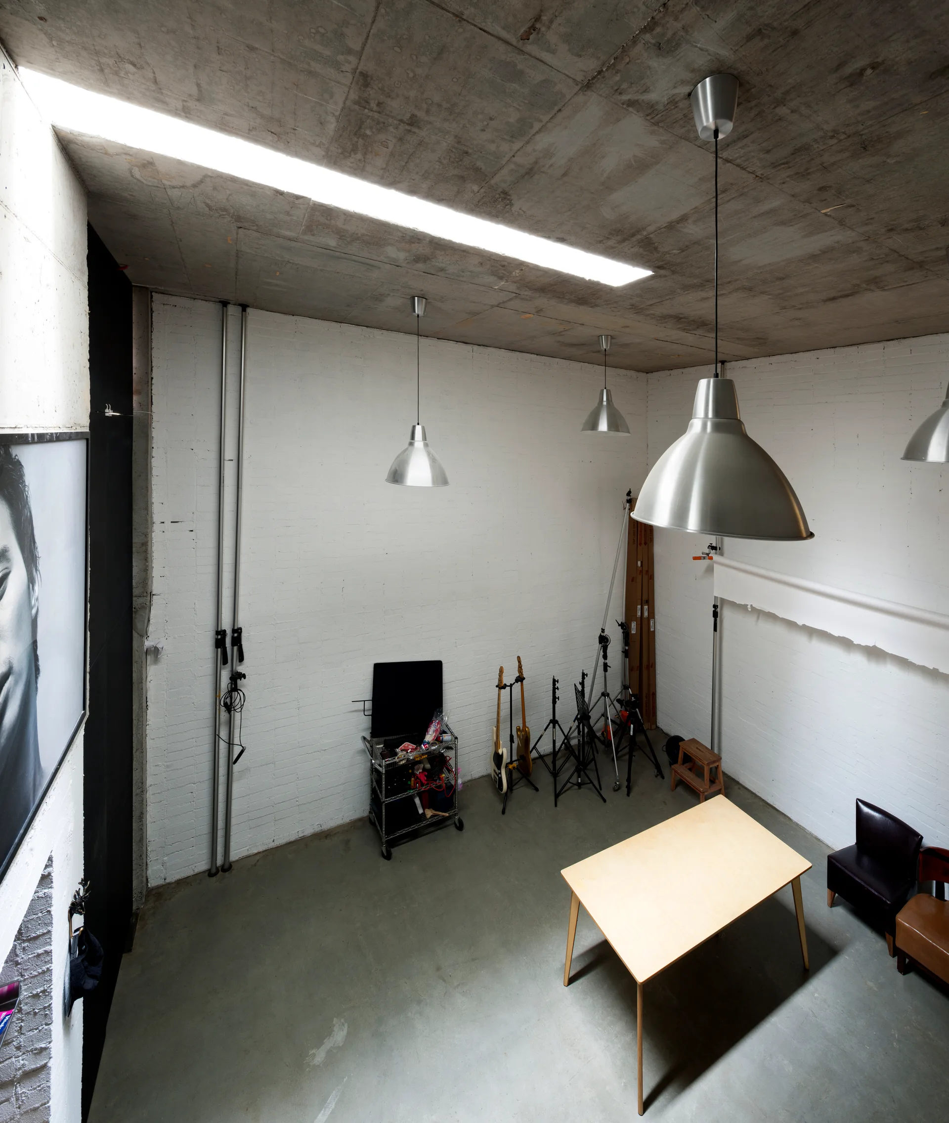 An industrial studio space featuring white painted brick walls, a raw concrete ceiling with silver pendant lights, and a light wood table.