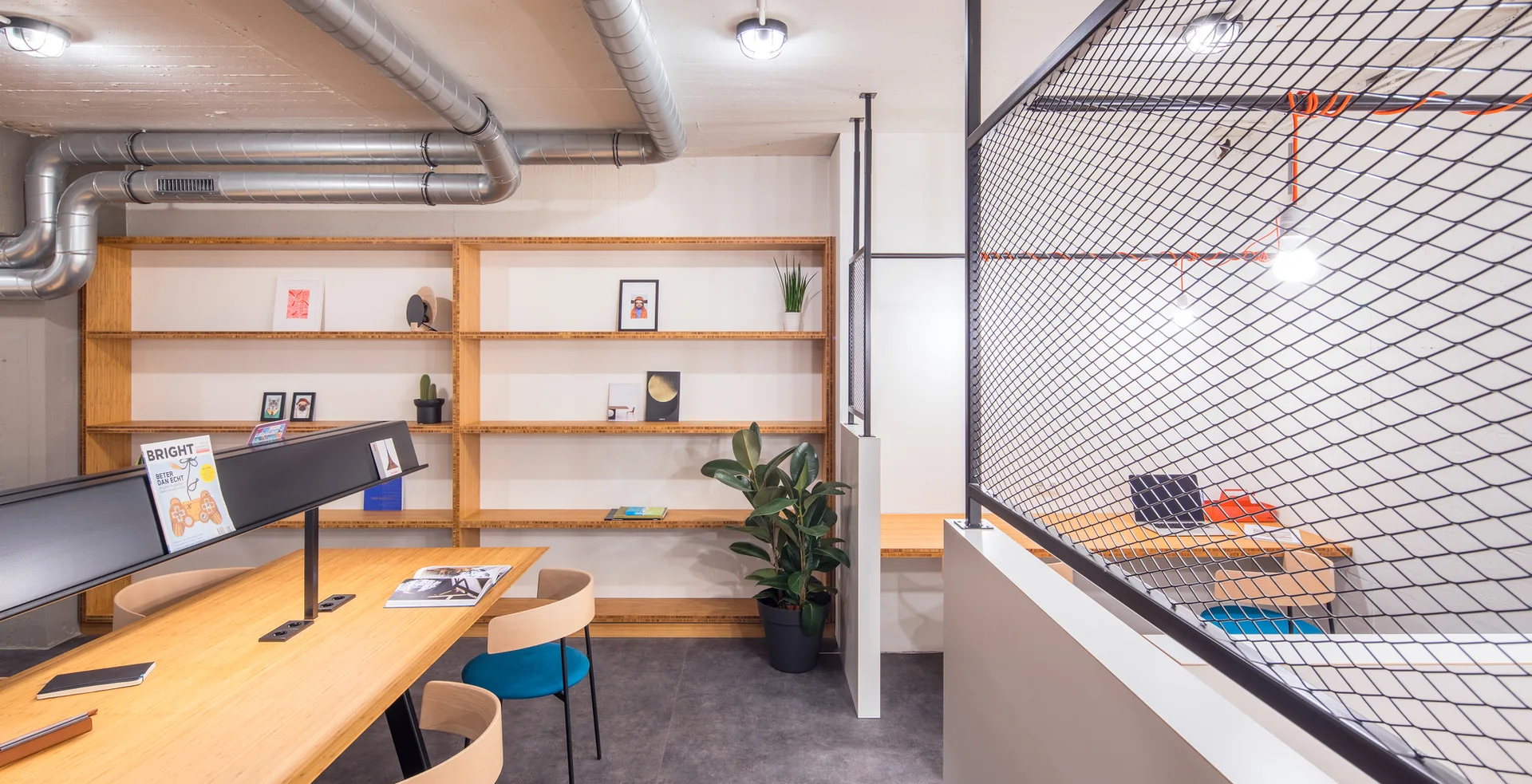 A modern office workspace features light wood desks, matching wall-mounted open shelves, exposed metal ducts, and a black wire mesh room divider.