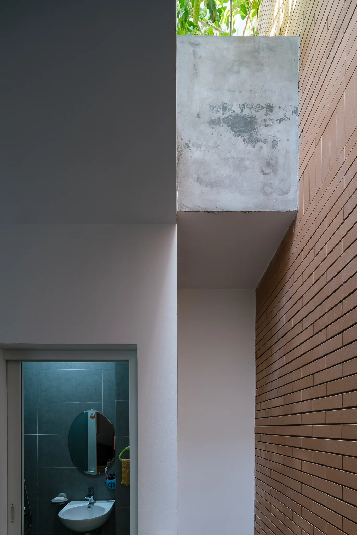 A viewpoint showing a bathroom with a white sink and round mirror framed by an outer hallway with a brown brick wall and raw concrete beam.