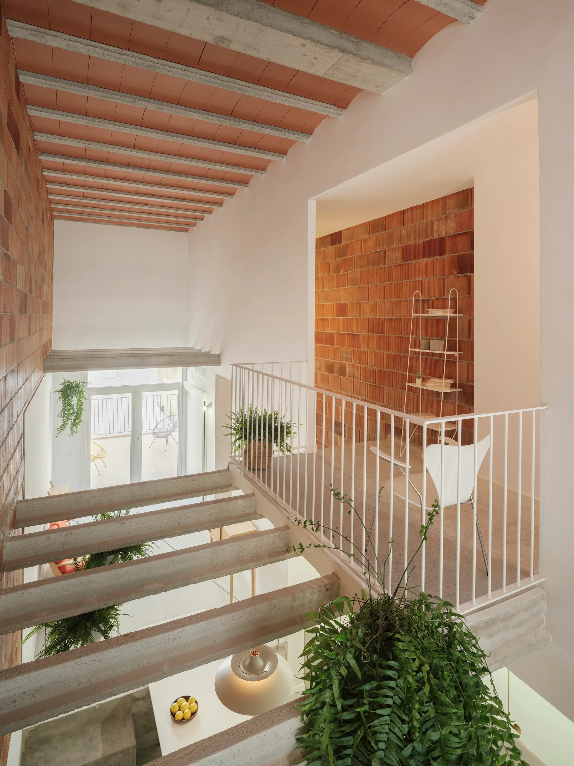 A multi-level interior features a floating concrete staircase, white metal railings on a landing with a white chair, and exposed brick walls.