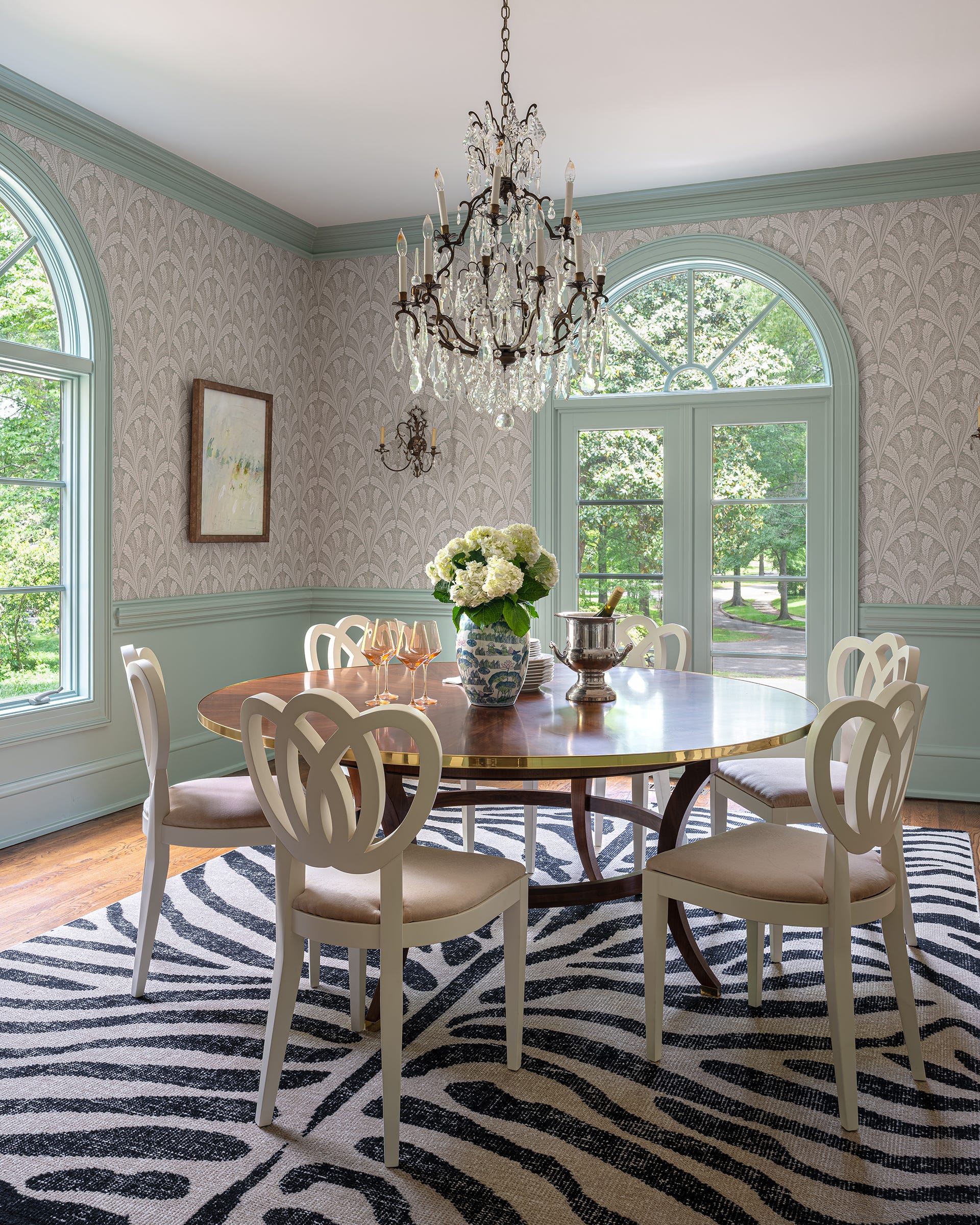 A round wooden dining table and white chairs sit on a black and white zebra print rug in a room with blue wainscoting and arched windows.